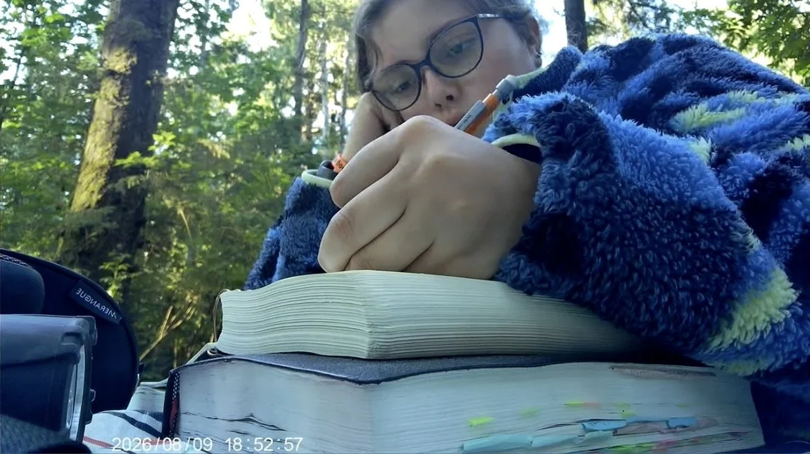Person with glasses writing in a notebook outdoors in a forest, surrounded by trees and green foliage.