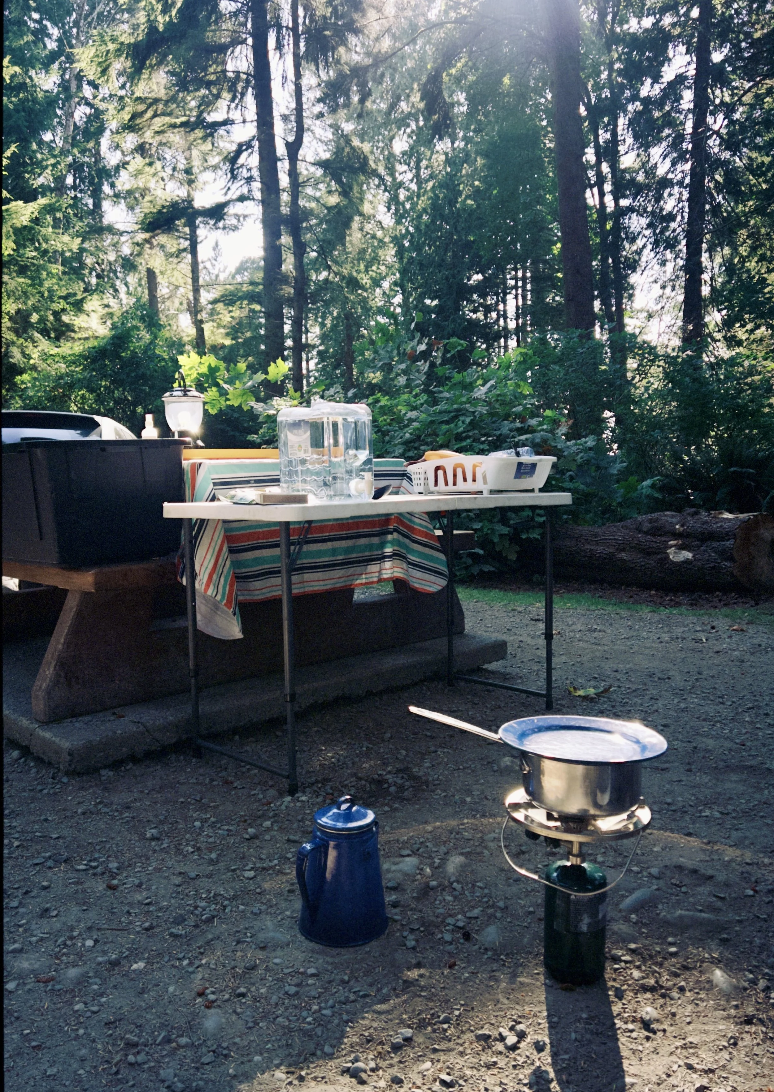 A camping scene with a table covered by a colorful striped cloth, placed outdoors in a wooded area with tall trees in the background. The table has a large pitcher of water and some plates. There is a small grill with a lid on the ground in front of the table, and a blue thermos and a small black container nearby. Sunlight filters through the trees.