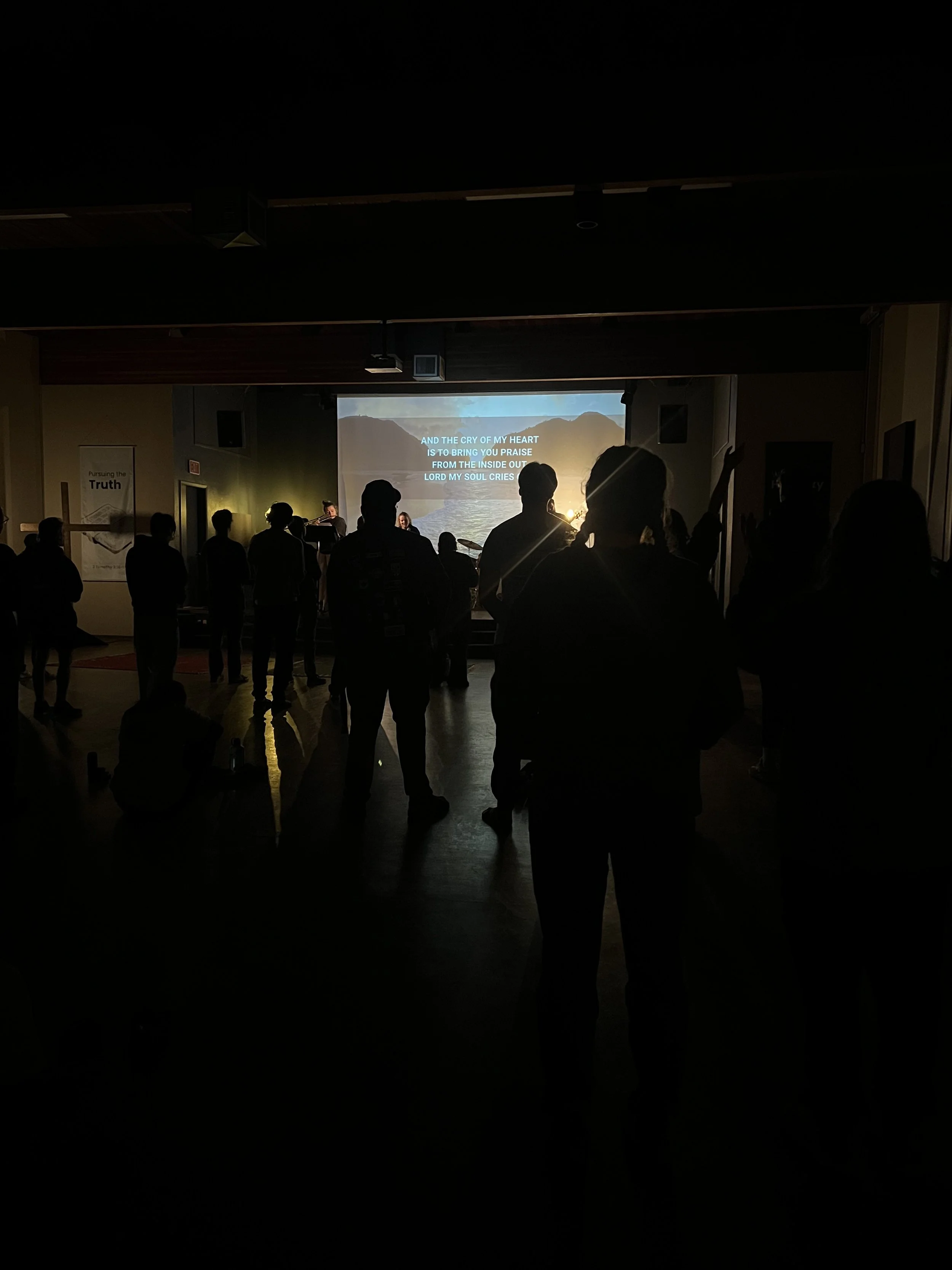 Silhouettes of people attending a concert or worship service in a dark room, with a large screen displaying lyrics over a mountainous landscape.