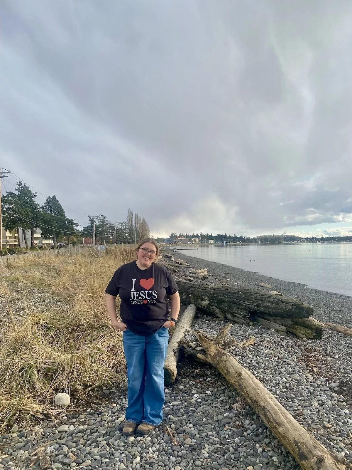 A woman standing on a rocky shoreline by the water, wearing a black T-shirt with red and white text that says 'I love Jesus' and blue jeans, smiling with her hands in her pockets, under a cloudy sky.
