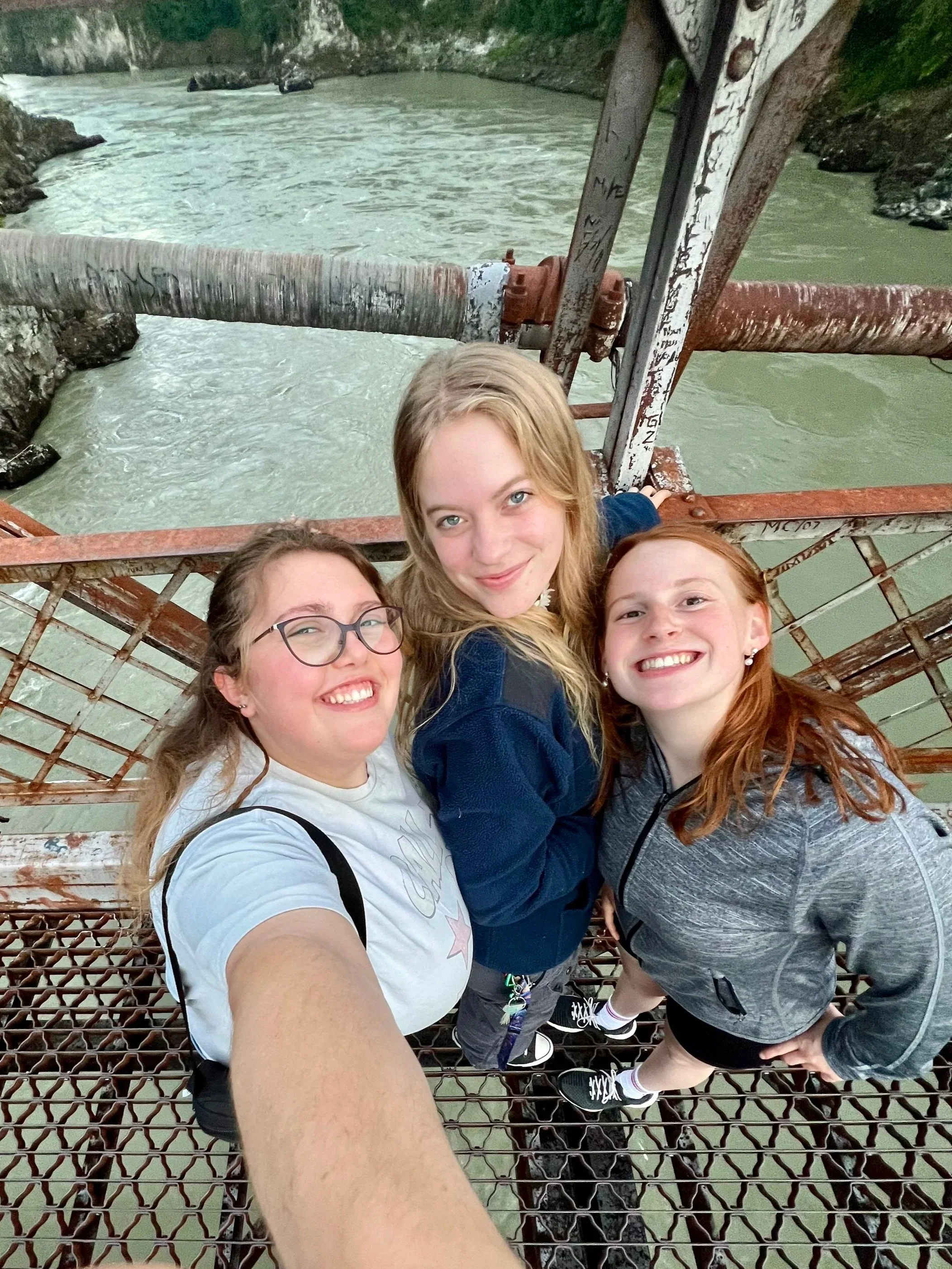 Four young women taking a selfie on a rusty metal bridge over a rushing river, with rocky walls and lush greenery in the background.