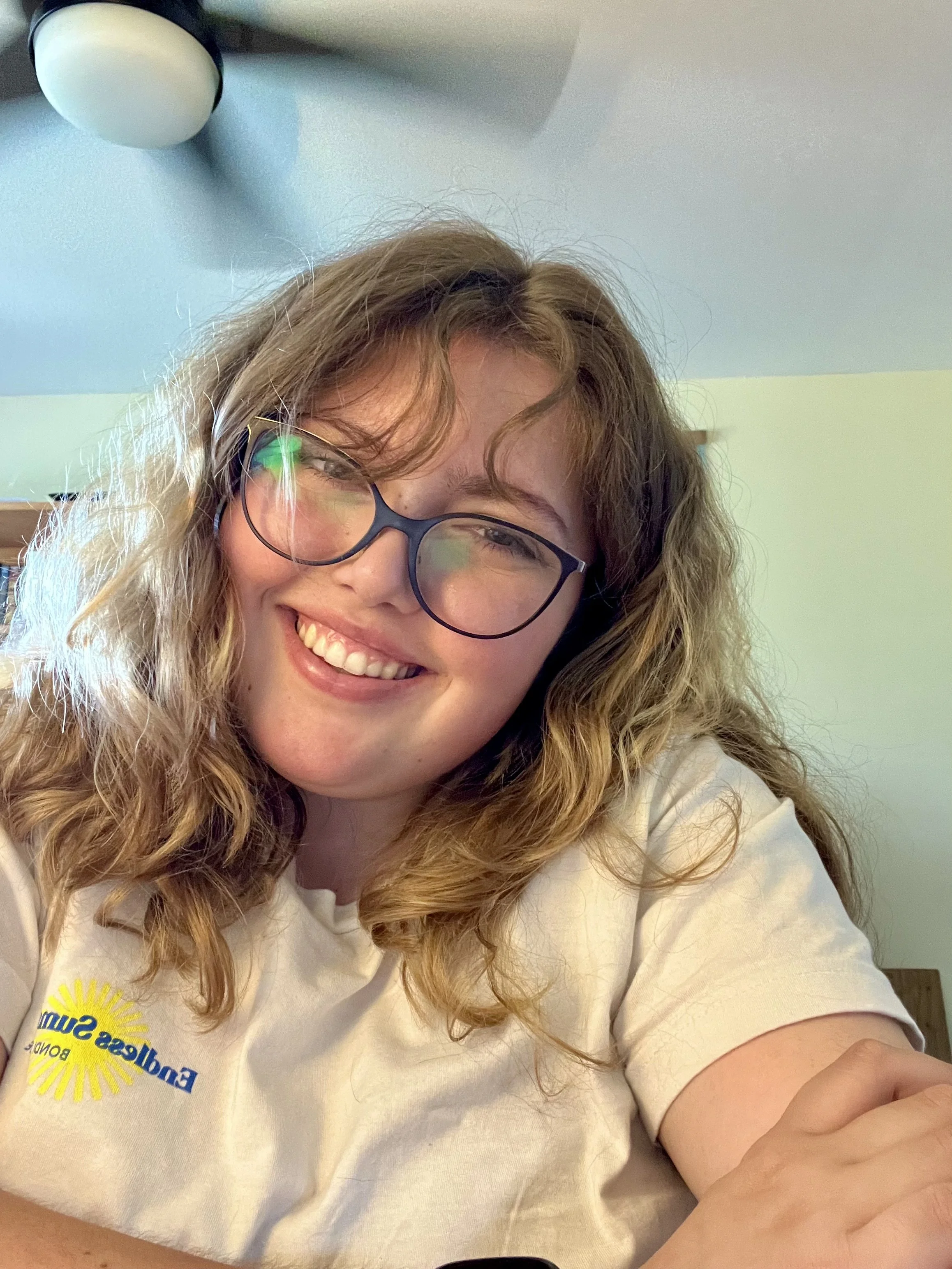 A smiling woman with curly hair and glasses wearing a light-colored T-shirt with a sunrise logo.