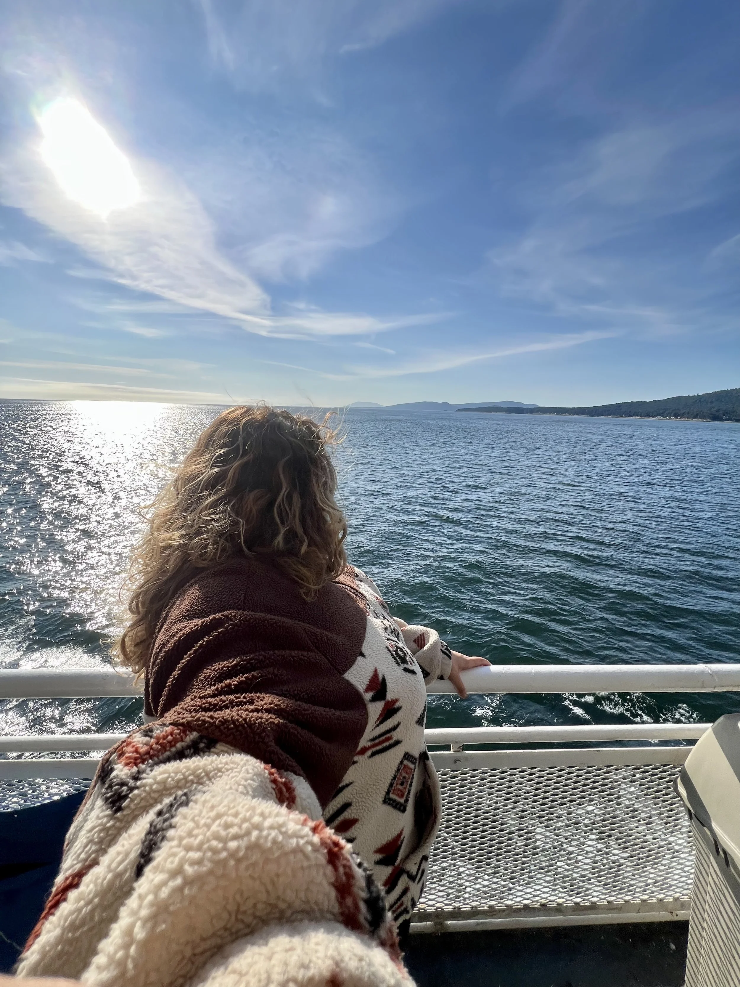 A woman with curly hair wearing a patterned sweater is sitting near a white railing on a boat, looking out at the ocean and distant landmasses under a clear, sunny sky.