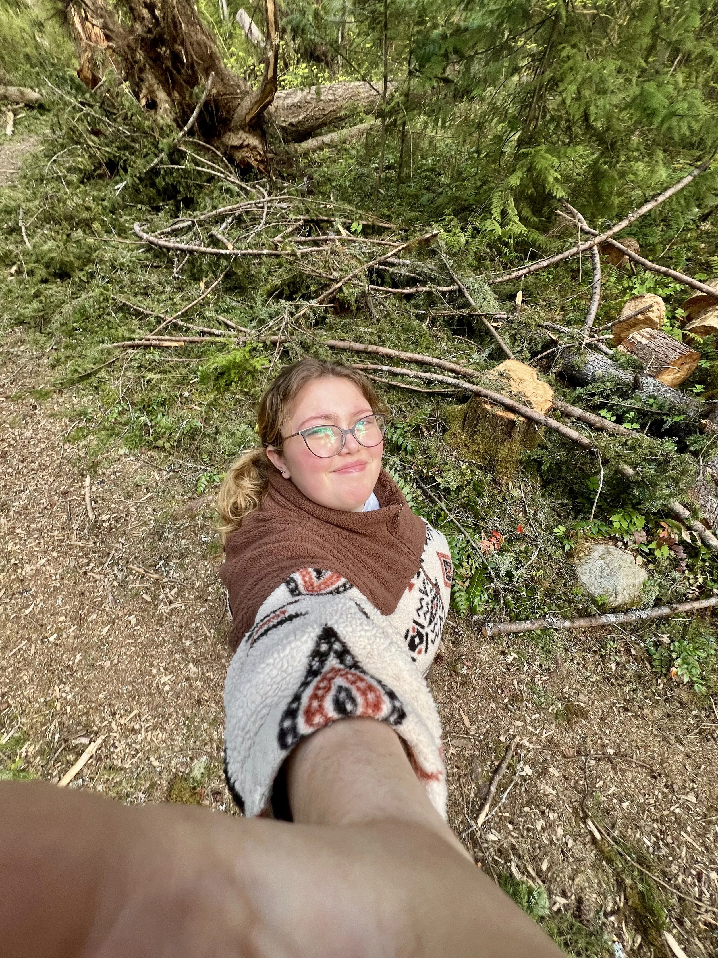 Young woman taking a selfie in a forest, with trees and fallen branches in the background.