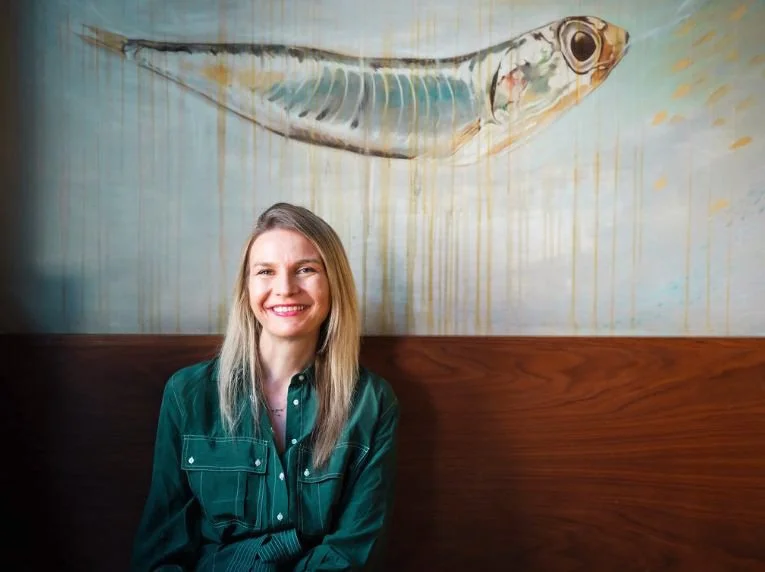 A woman with long blonde hair smiling while sitting in front of a mural of a fish painted on the wall.