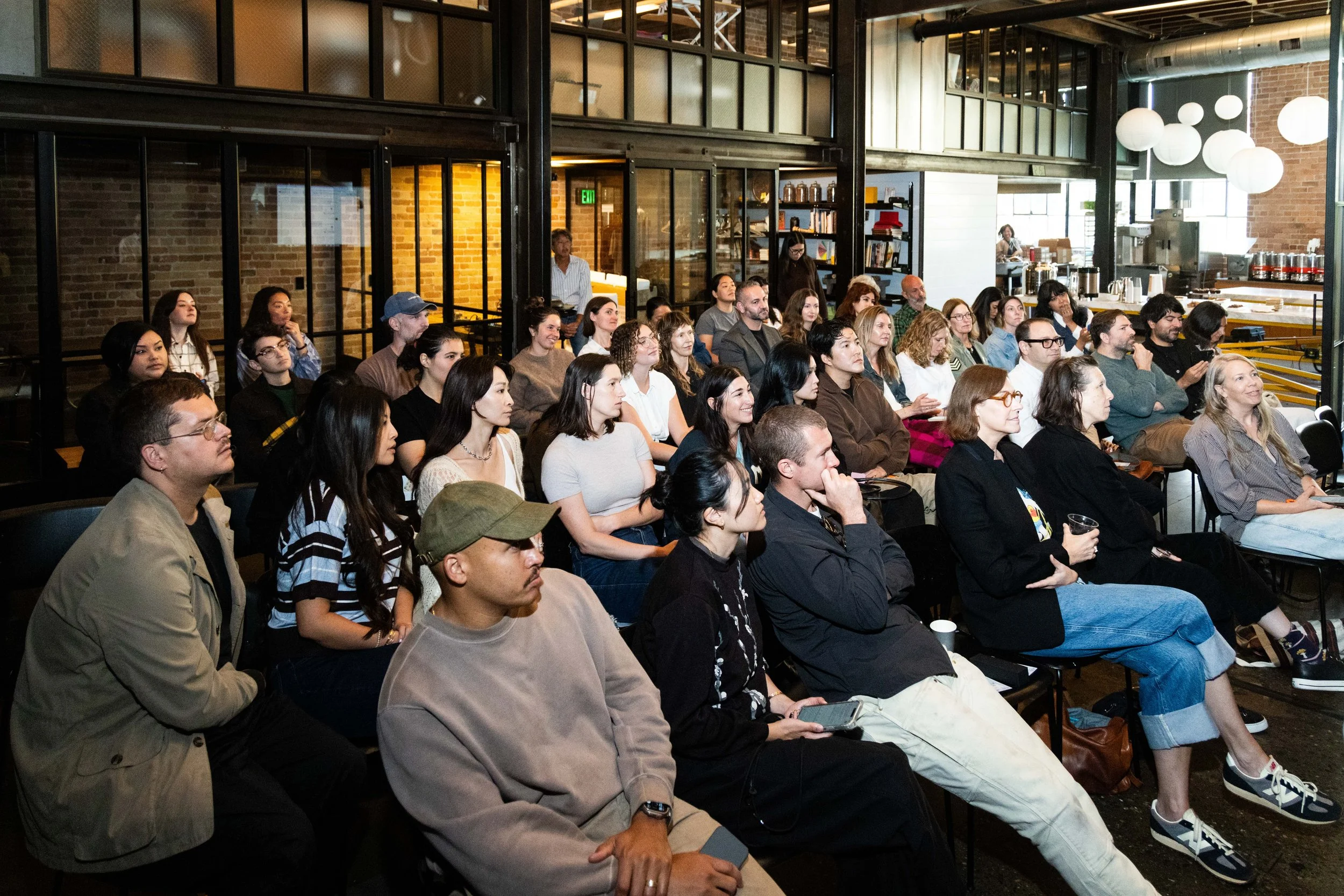 Audience attending a presentation or event inside a modern industrial-style venue with exposed brick walls and large windows, with some people taking notes and listening attentively.