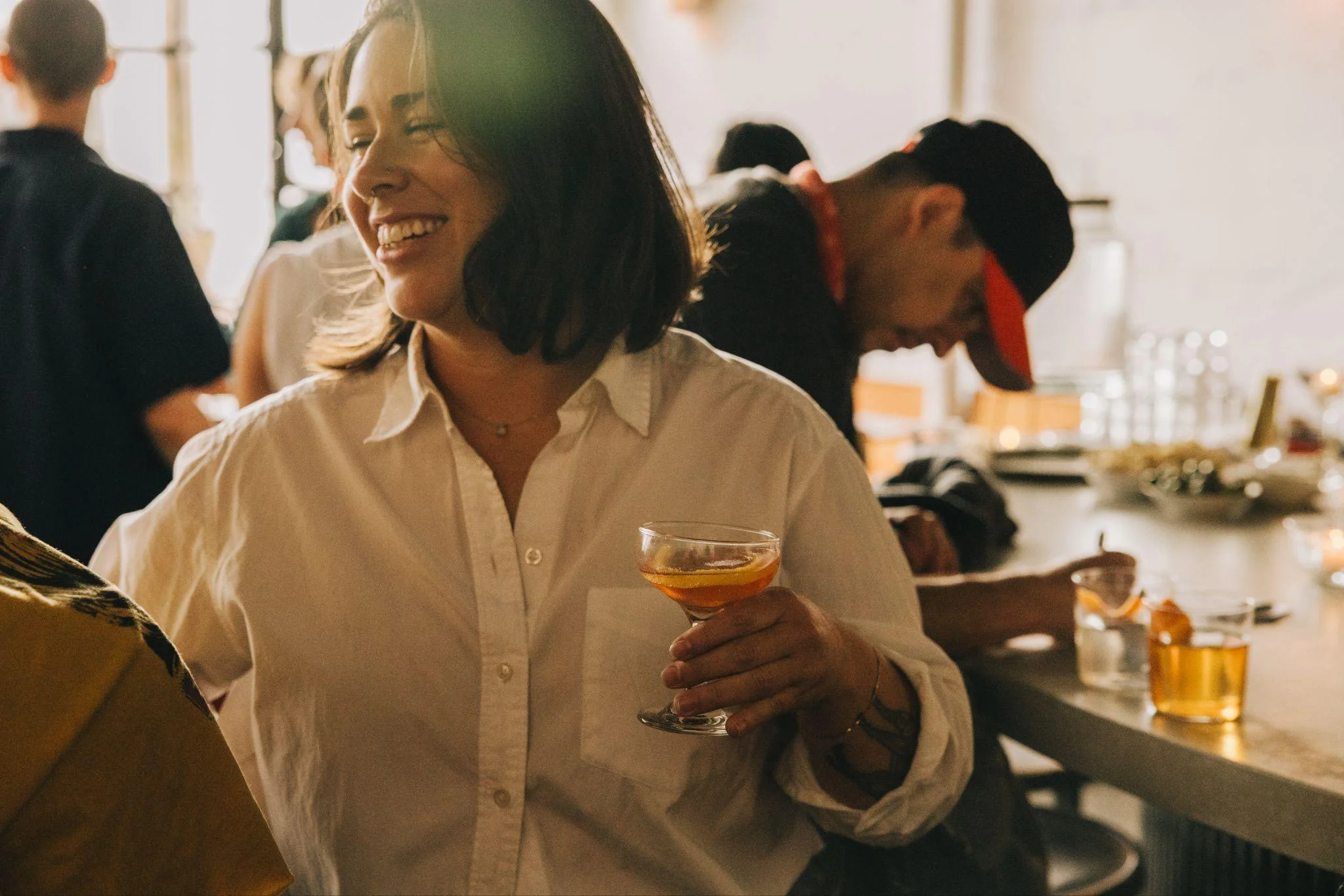 A woman with shoulder-length dark hair wearing a white shirt is holding a cocktail glass and smiling at a social gathering. Other people are in the background, and the setting appears to be indoors with warm, natural lighting.