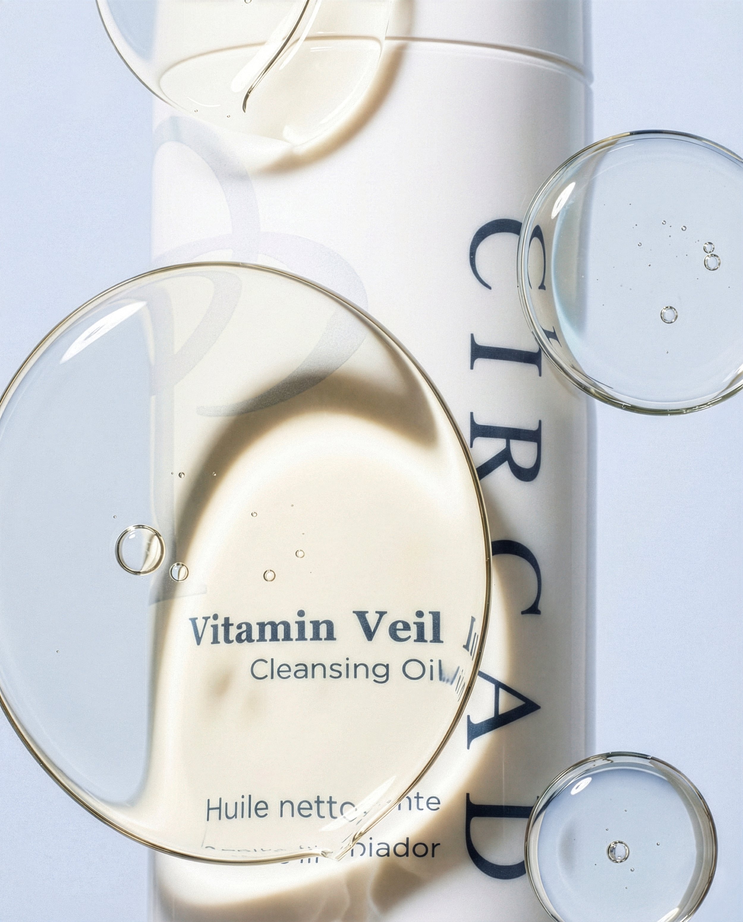 Close-up of a white box with the text 'Vitamin Veil Cleansing Oil' partially visible. Two clear glass bowls with some oil and bubbles sit on top of or around the box. The background includes printed text, with some letters visible.