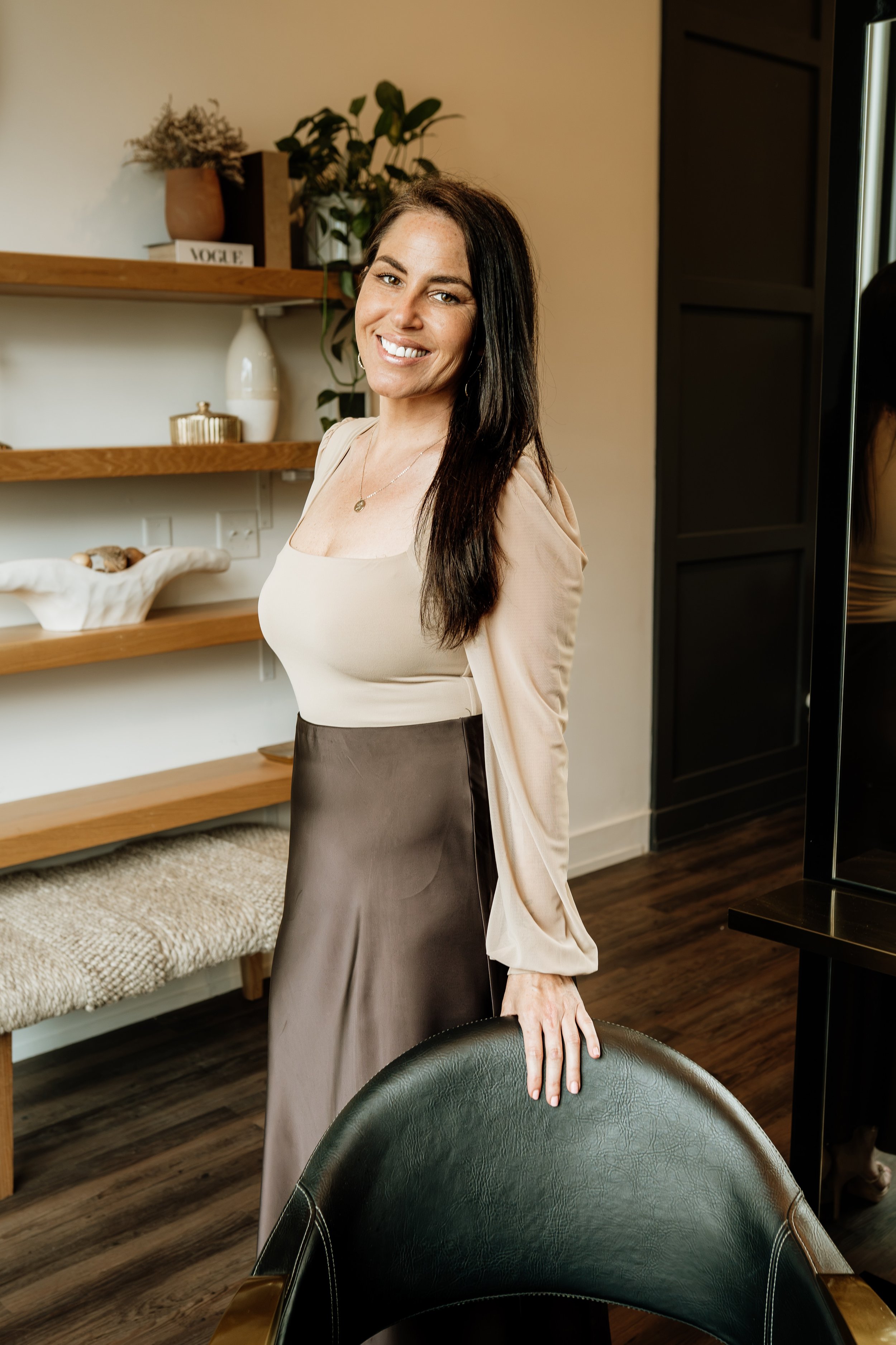 A woman with dark hair smiling and standing in a room with wooden shelves, plants, and decorative items. She is dressed in a beige top and a satin skirt, resting her hand on a black leather chair.