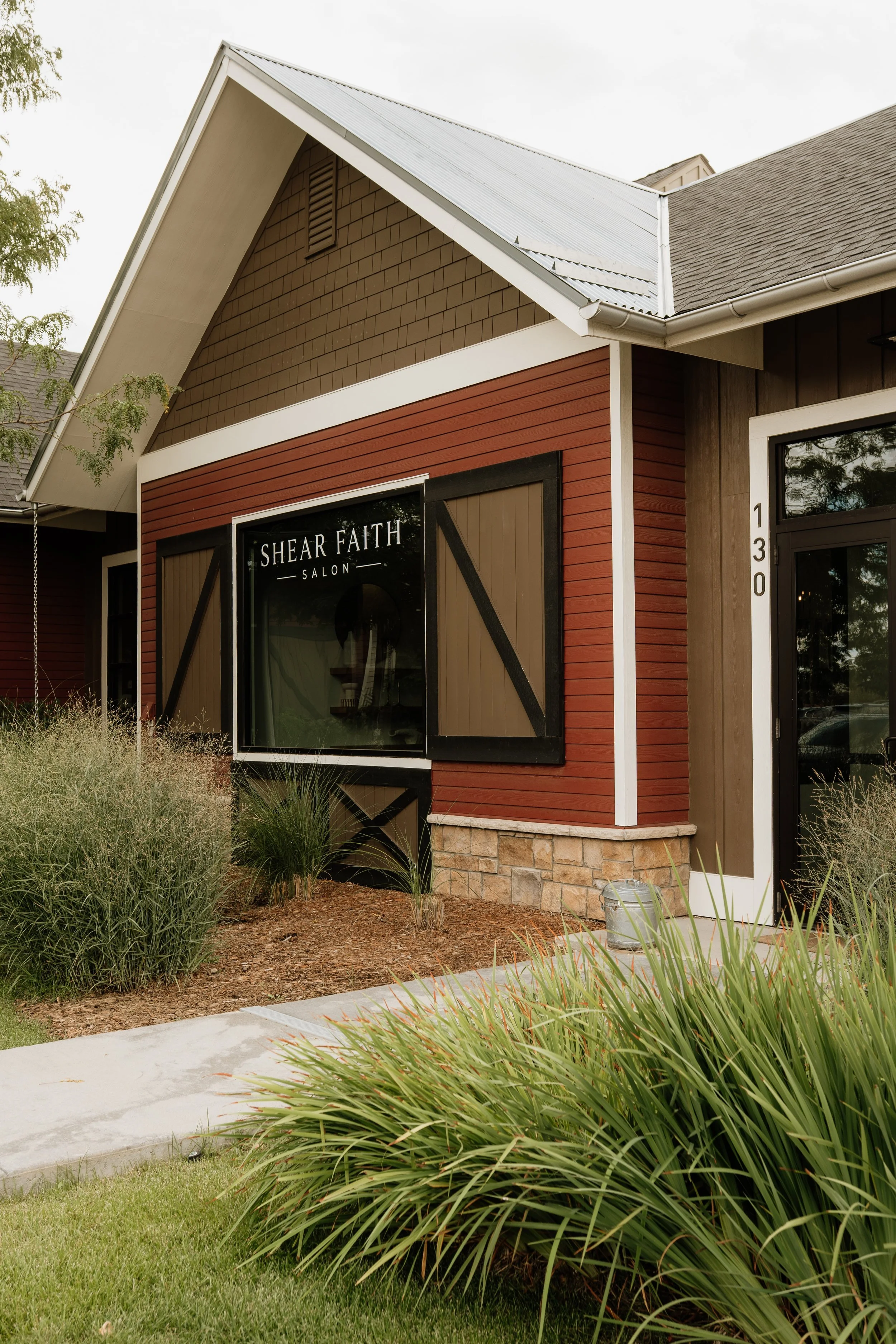 Exterior view of a modern house with a red wooden siding, large front window with black trim, and a stone foundation. The house has a gabled roof with gray shingles and a metal roof section. There are plants and grass in the yard, and a concrete walkway leads to the entrance.