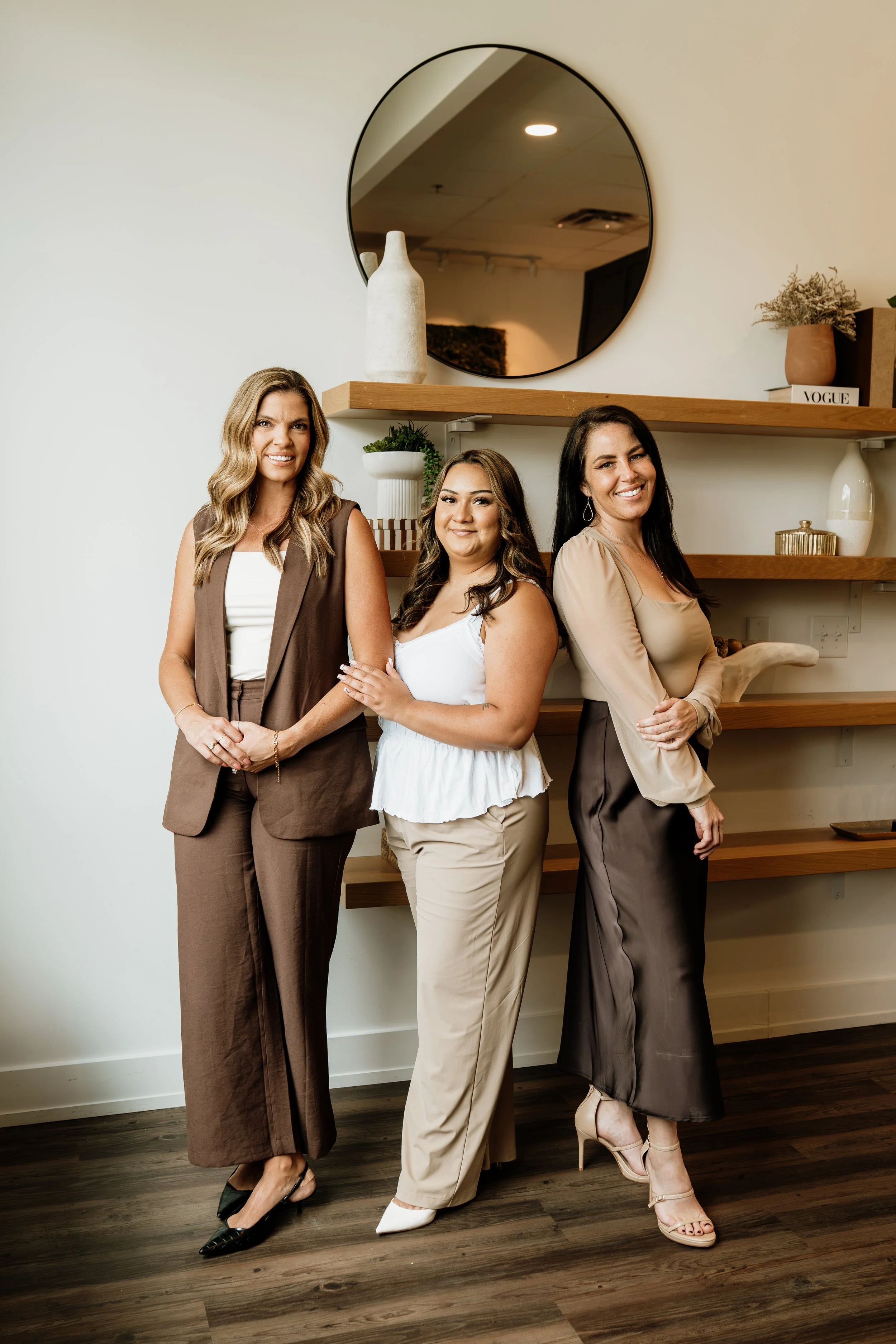 Three women standing next to each other indoors, smiling, in front of a wooden shelf with decorative items and a round mirror on the wall.