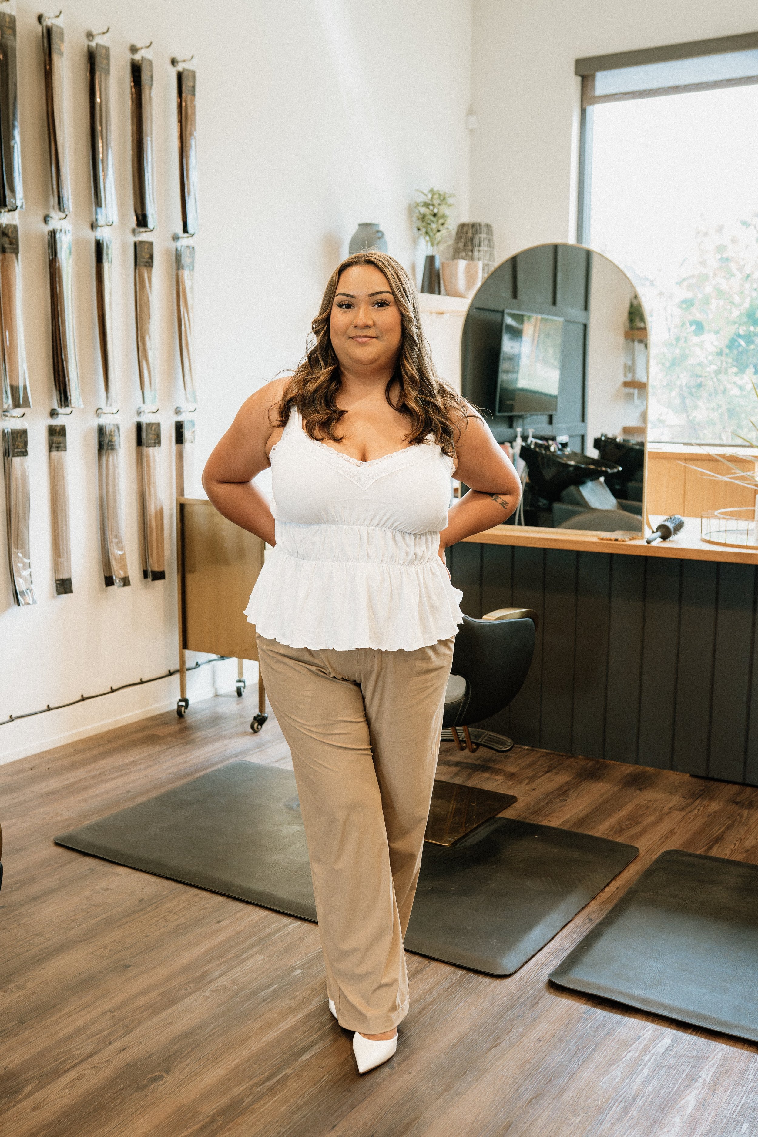Woman with long wavy hair wearing a white sleeveless top, beige pants, and white shoes standing in a modern salon with hair extensions displayed on the wall, a large mirror, and salon equipment visible.