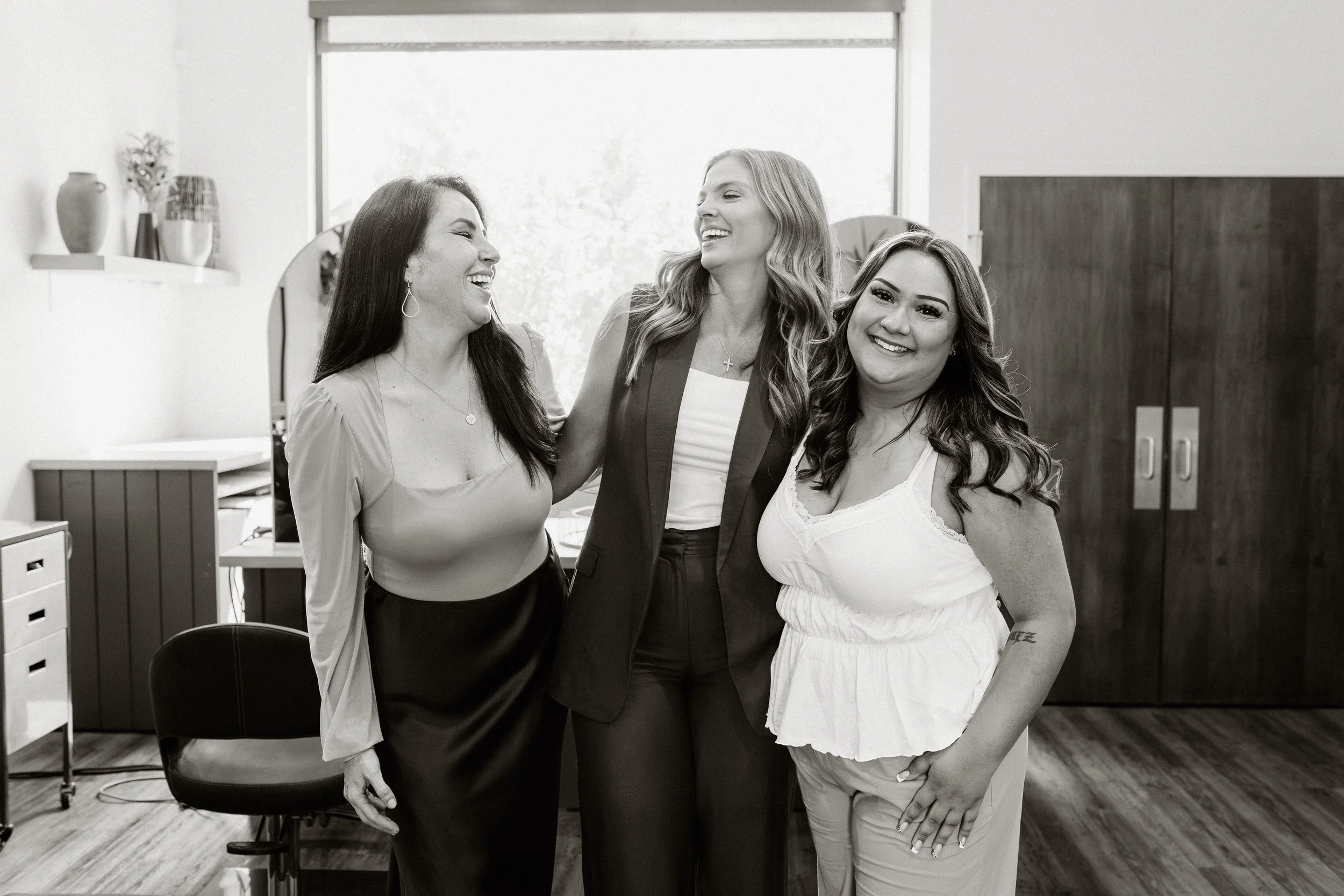 Three women smiling and laughing together in an indoor setting, with decorative vases on shelves and a large window in the background.