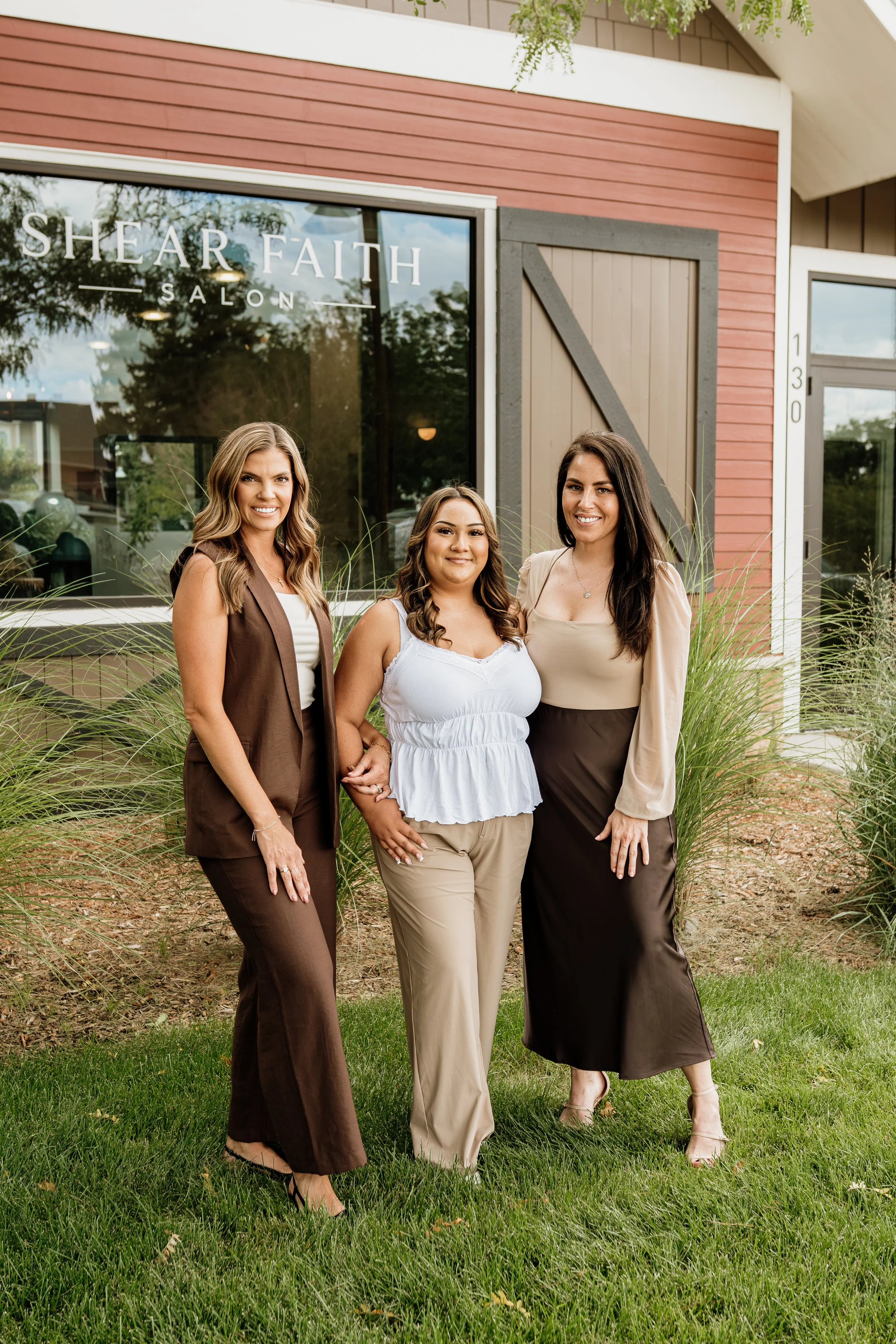 Three women standing outdoors in front of a building with large glass window and wooden accents. The sign on the window reads 'SHEAR FAITH SALON'. The women are dressed in business casual attire, smiling, and posing for the camera. There are green plants and grass in the foreground.