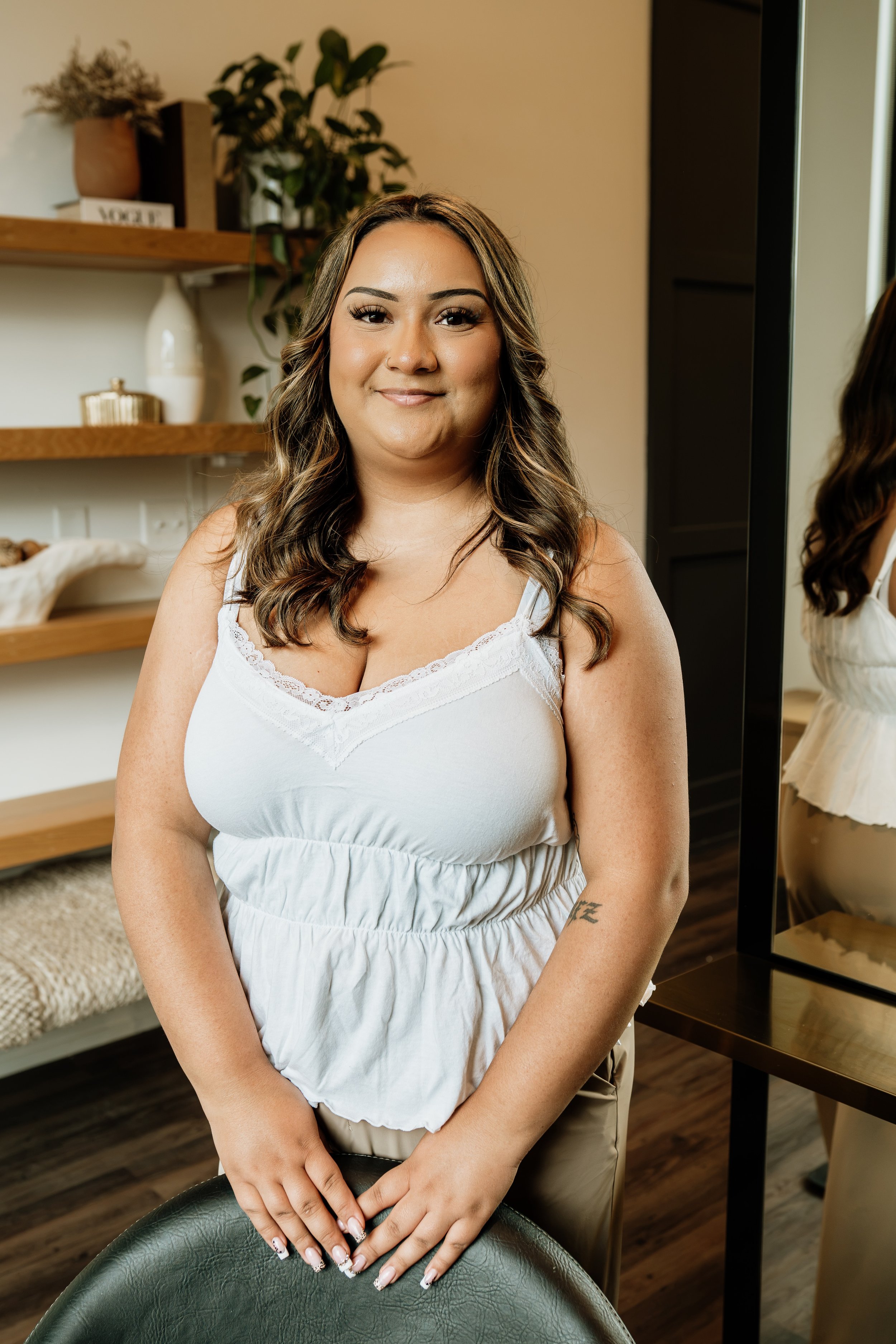 A woman with wavy brown hair and makeup, wearing a white sleeveless top with lace trim, standing in a room with wooden shelves, plants, and a mirror.