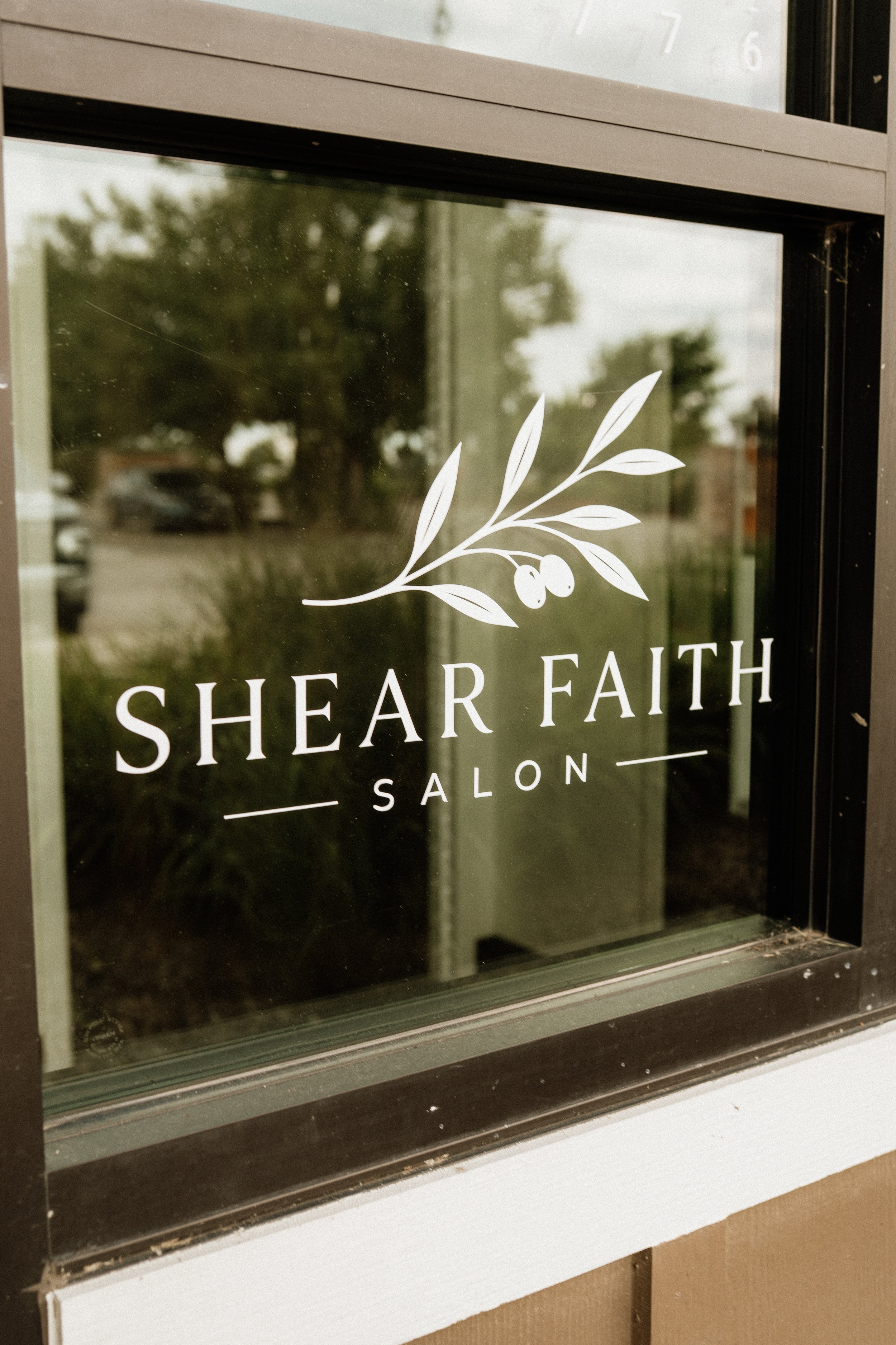 A glass door or window with a sticker that reads "Shear Faith Salon" and has a decorative branch with leaves and berries design.