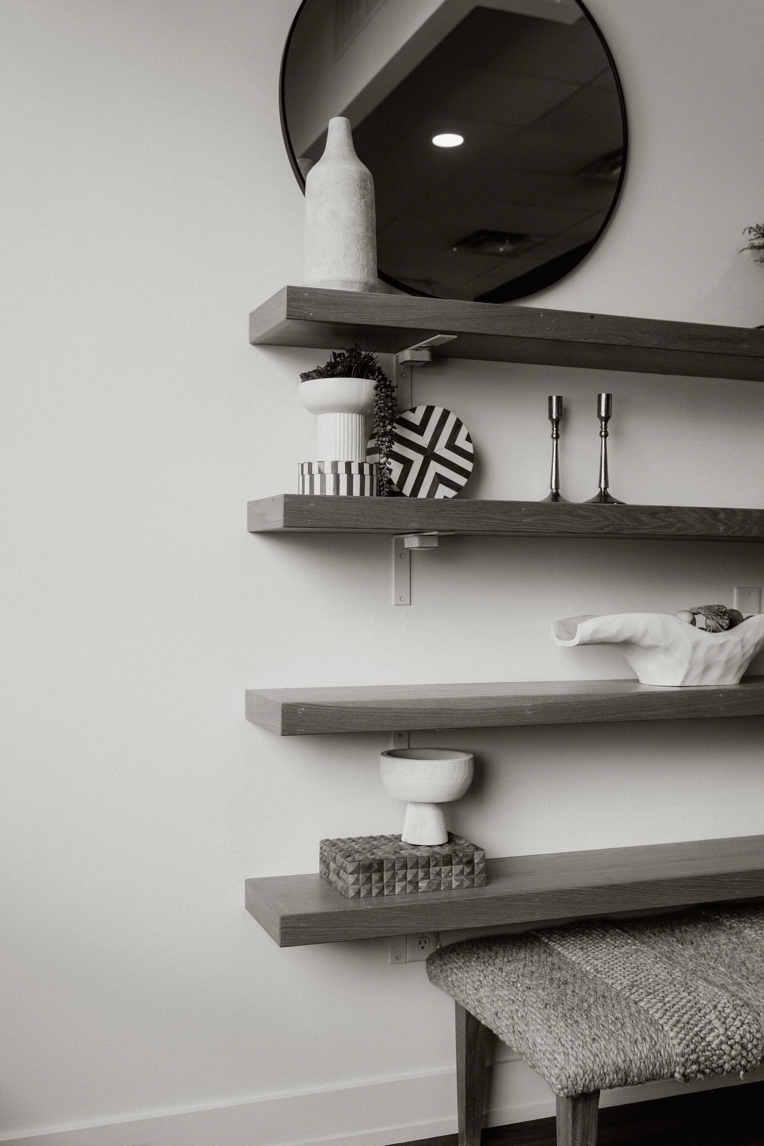 Wall-mounted wooden shelves displaying decorative objects, a round mirror, and a chair with a textured blanket draped over it.