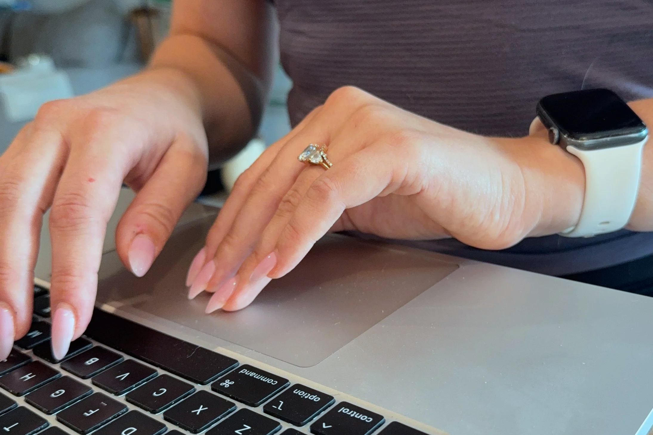 Close-up view of a person typing on a laptop keyboard with their left hand, wearing a white smartwatch and a ring with a large gemstone on the ring finger.