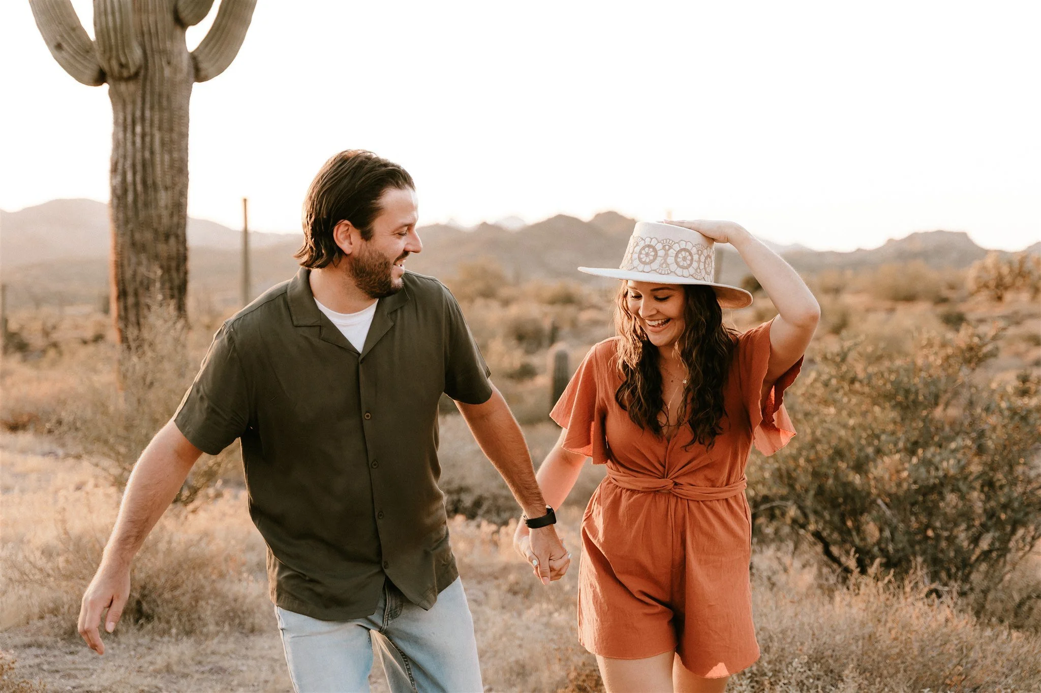 A man and woman holding hands and smiling in a desert landscape with a cactus and mountains in the background, during sunset.