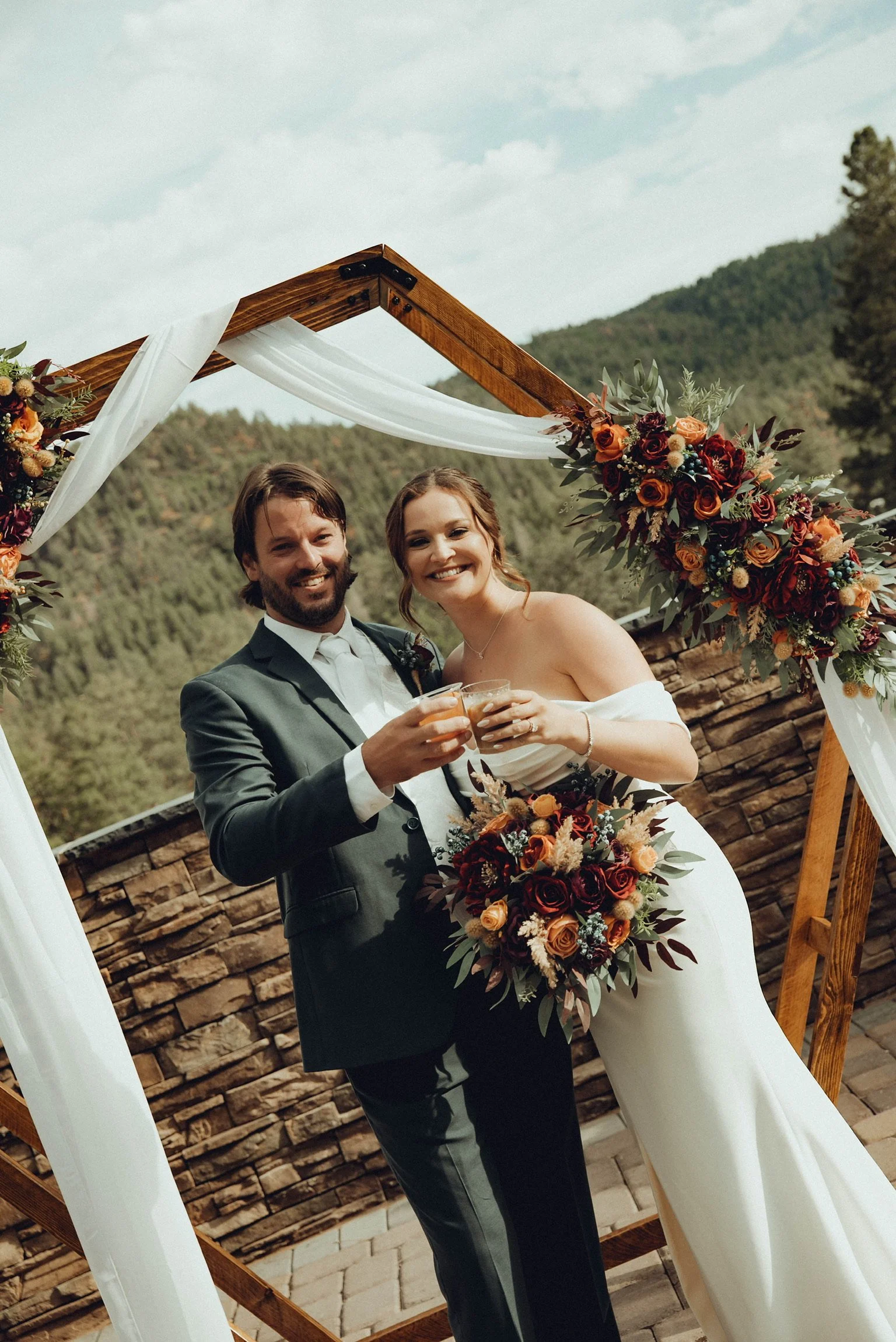 Happy bride and groom holding drinks under a wooden wedding arch decorated with flowers and greenery at an outdoor wedding ceremony with mountain scenery in the background.