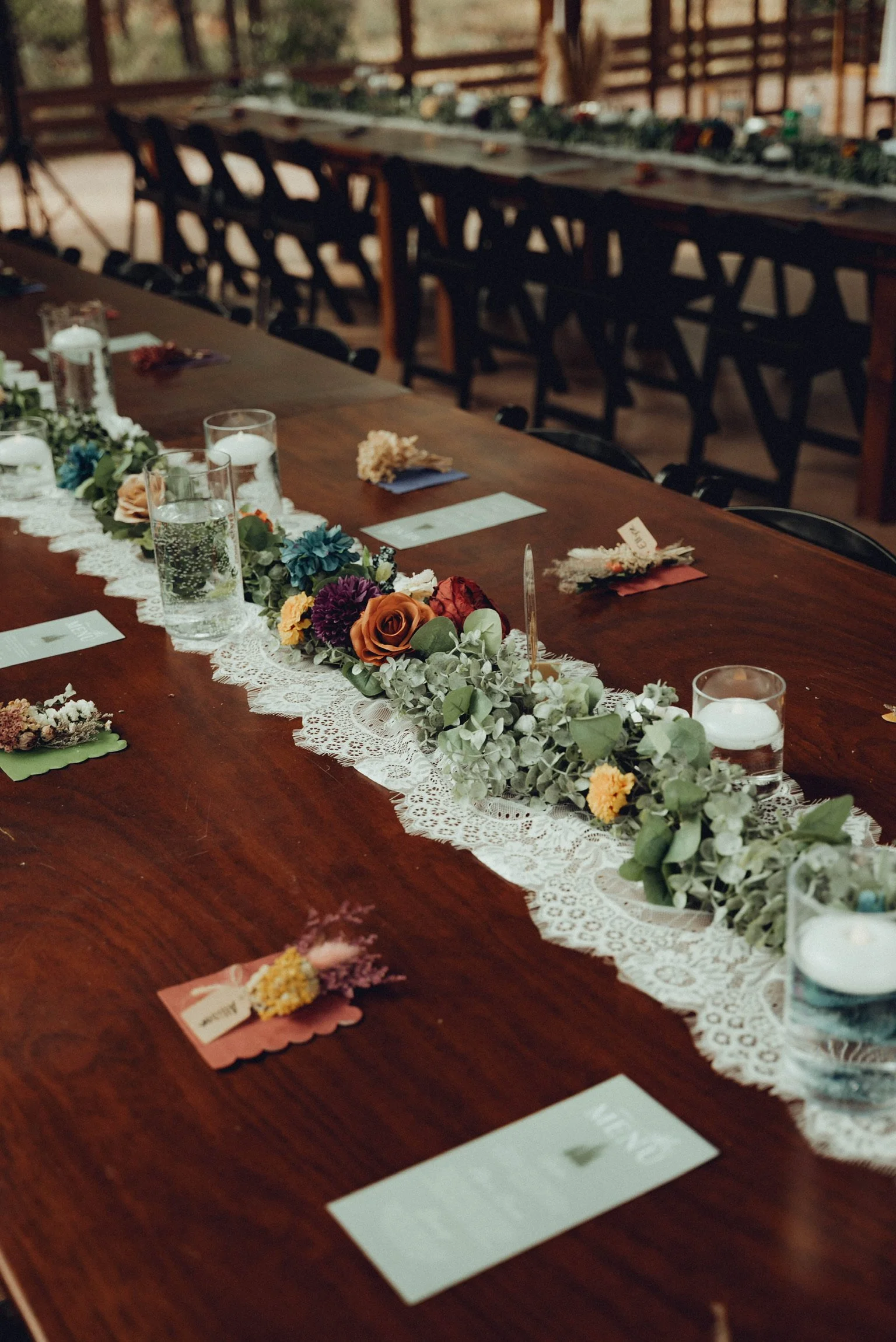 A long wooden table decorated with a lace runner, floral arrangements, candles, and place cards for an event in a rustic, barn-style setting.
