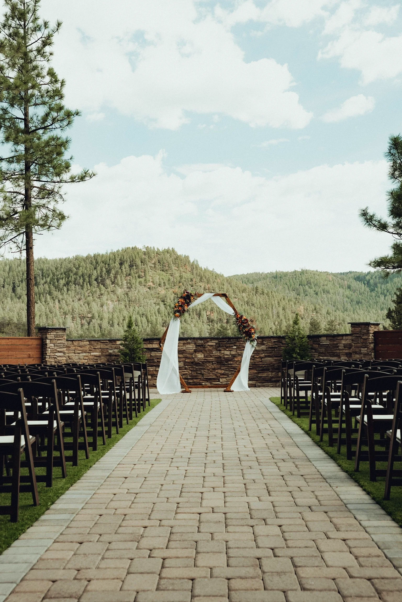 Outdoor wedding setup with black chairs arranged on either side of a brick pathway, leading to a wooden arch decorated with flowers and white fabric, surrounded by trees and mountains under a partly cloudy sky.