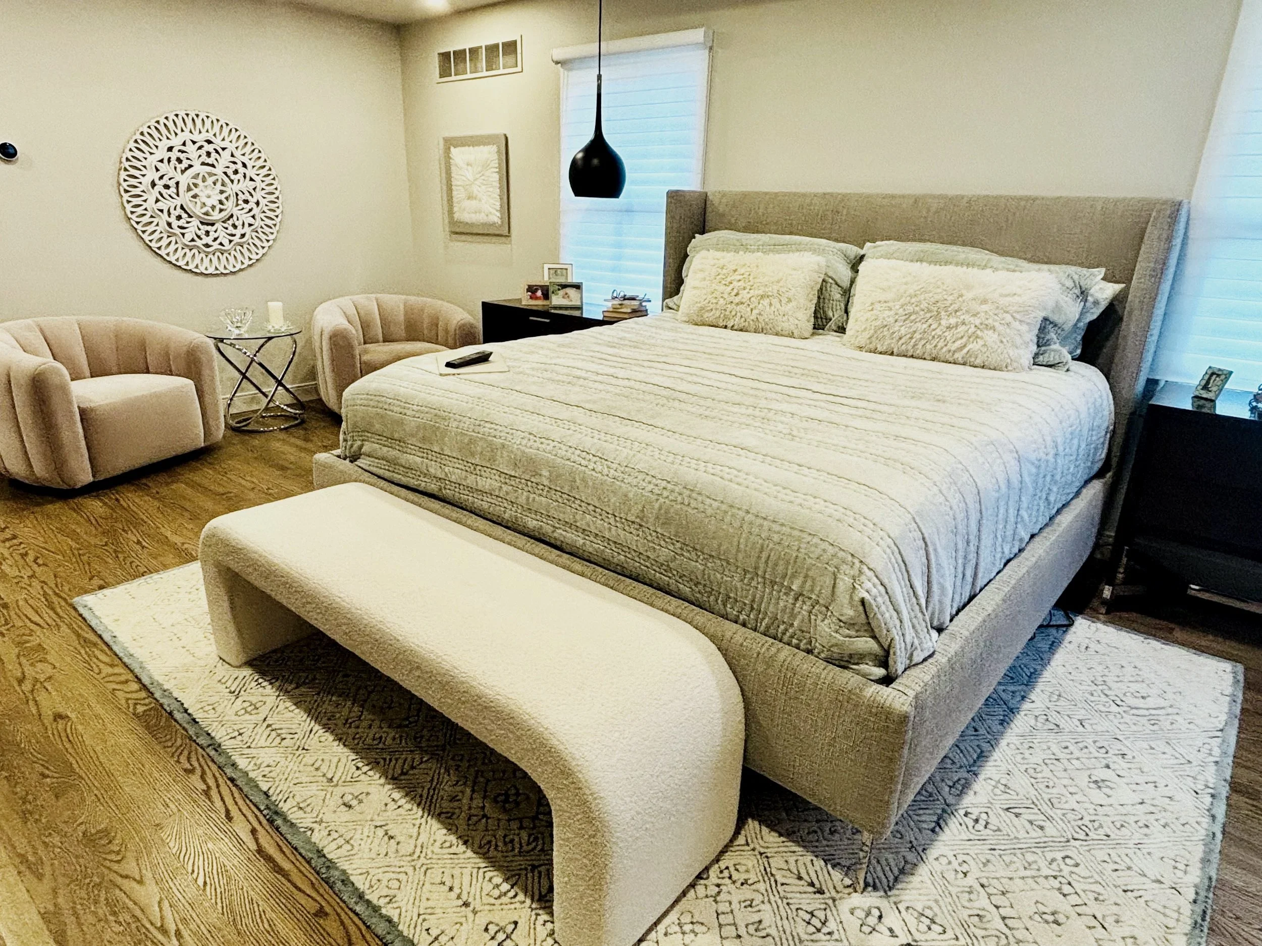 A bedroom with a large bed with beige bedding and multiple pillows, a nightstand with framed photos and books, two beige armchairs, a small round table with a glass bowl and a candle, a large decorative white wall art, and a window with white blinds and a black hanging light fixture.