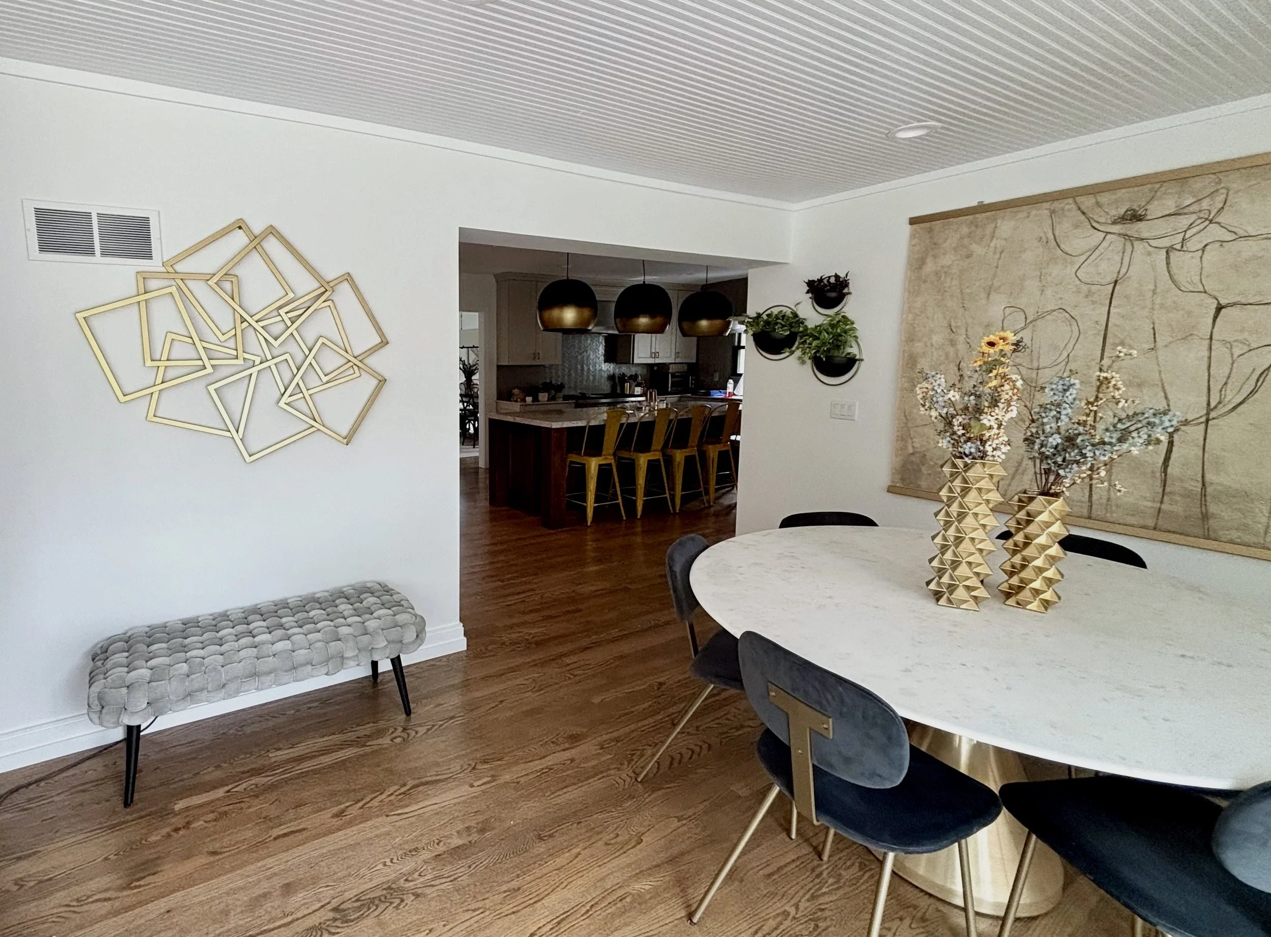Modern dining room with a white round table, dark blue chairs, gold and geometric decor, a wall-mounted gold metal art piece, a floral arrangement in a geometric gold vase, and a large floral artwork on the wall. In the background, there is a kitchen with three pendant lights over a counter and barstools.