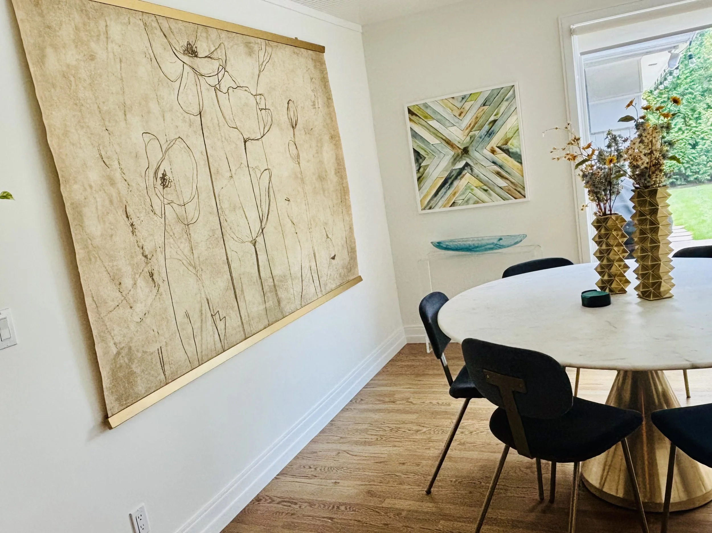 A dining room with a large beige and brown throw blanket hanging on the wall, a round wooden table with black and tan chairs, two vases of dried flowers on the table, a window with a view of greenery, and framed artwork on the wall.
