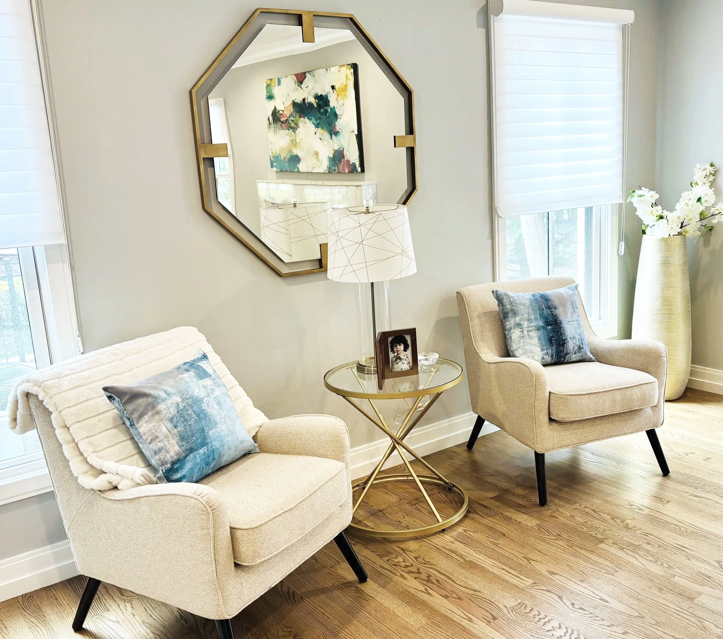 Living room corner with two beige armchairs with decorative pillows, a glass top side table with a framed photograph, white curtains on windows, a large fancy mirror, a white lamp, and a vase with white flowers.