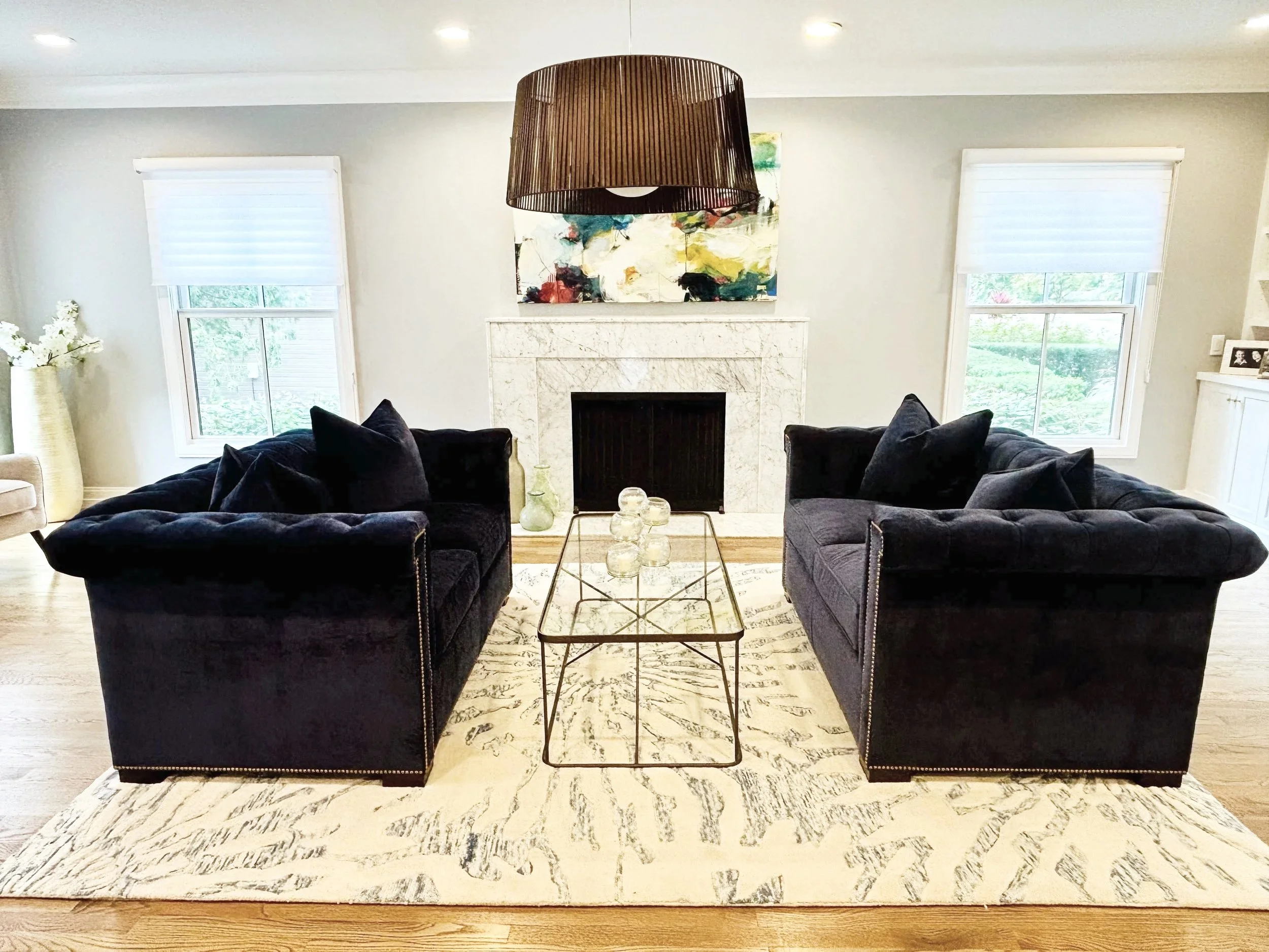 Living room with two black velvet armchairs facing each other around a glass-top coffee table, white marble fireplace with abstract art above, two windows with white blinds, and a cream-colored vase with white flowers on the left.