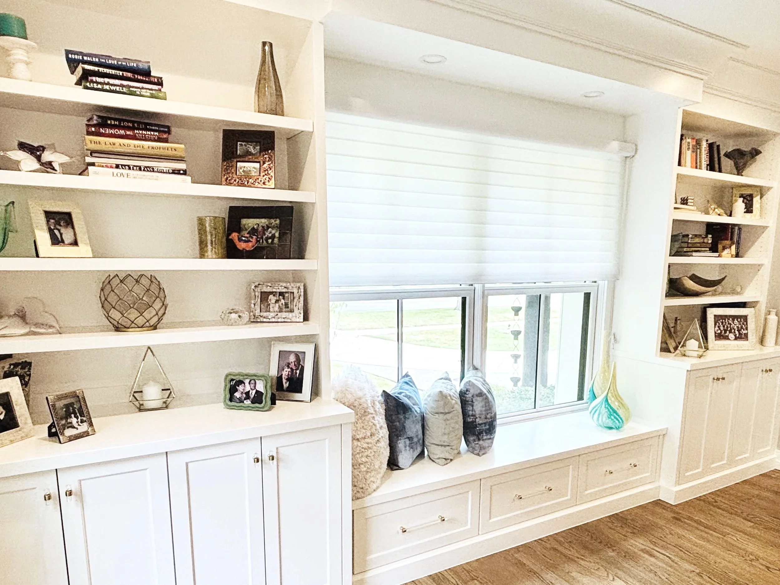 Built-in white bookshelf with decorative items, framed photographs, and family portraits, located in a living space near a window with blinds.