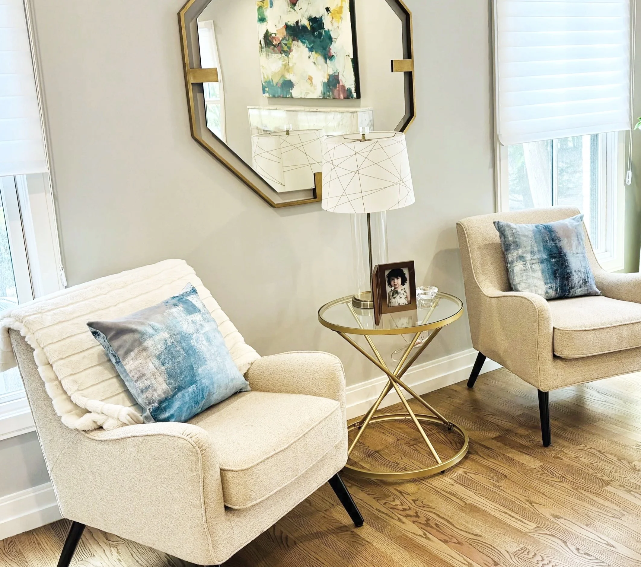 Living room corner with an off-white armchair, blue and white throw pillows, a gold side table with a framed photo and glass dish, a white table lamp with a geometric pattern, a large gold and black framed mirror, a colorful abstract painting, a beige chair with a blue pillow, and windows with white blinds.