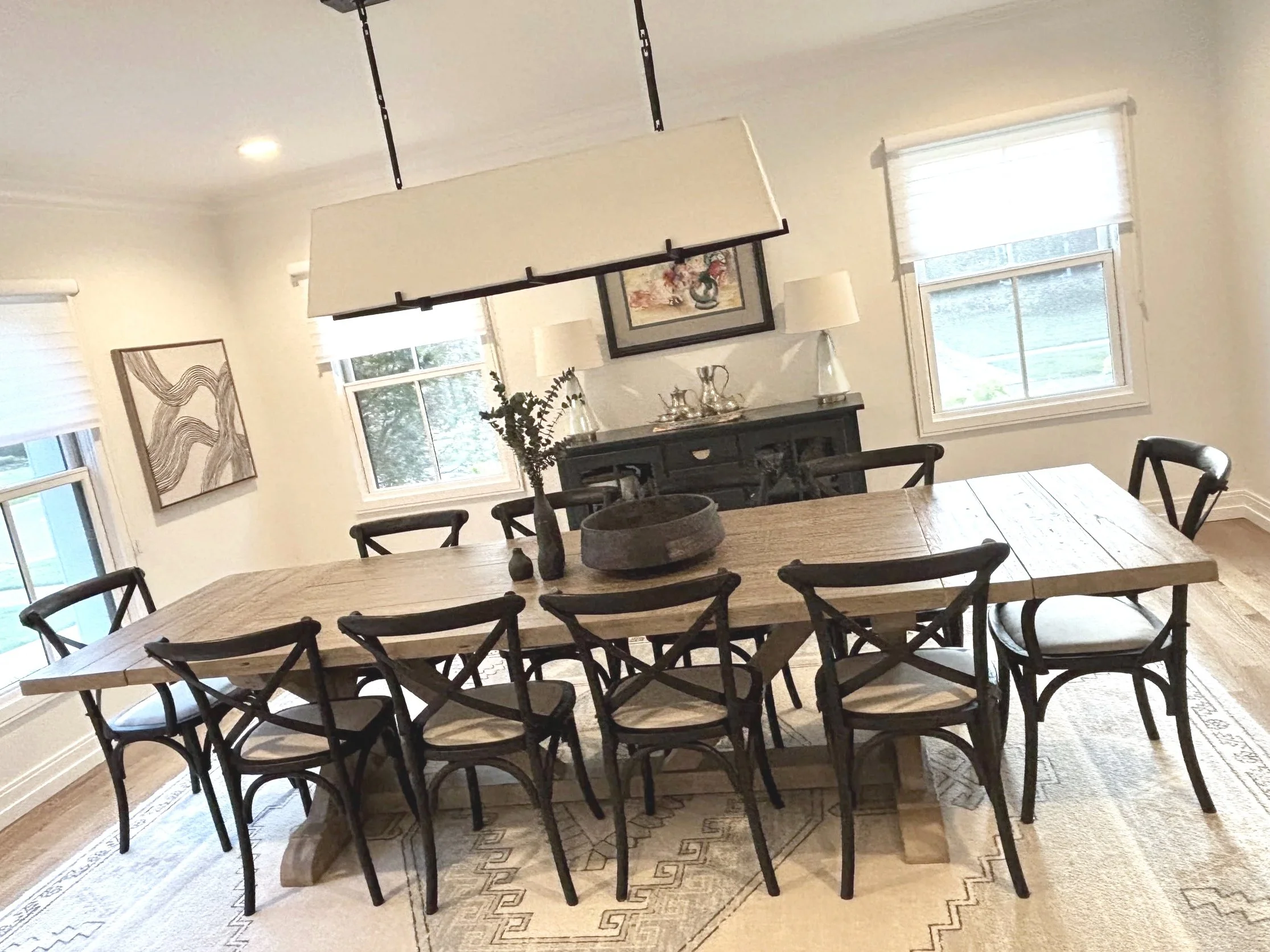 A dining room with a large wooden table surrounded by black wooden chairs, a black sideboard, and framed artwork on the cream-colored walls. There are two windows with white blinds, and a decorative lamp hanging from the ceiling.
