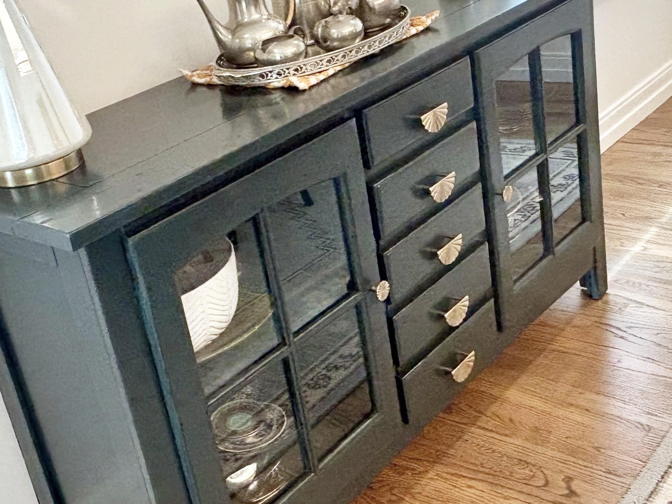 A black wooden sideboard with six glass-fronted drawers featuring gold fan-shaped handles. On top, there is a decorative tray with silver teapots and a lamp with a textured base.