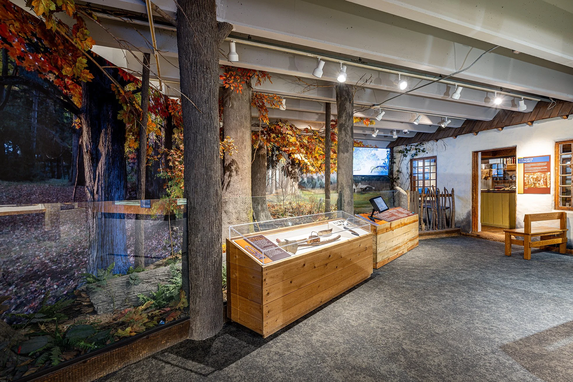 The Origins exhibit at the Detroit Historical Society. Indoor exhibit with artificial trees and autumn leaves, display cases with artifacts, informational signs, and historical decor resembling a rustic farm or pioneer setting.