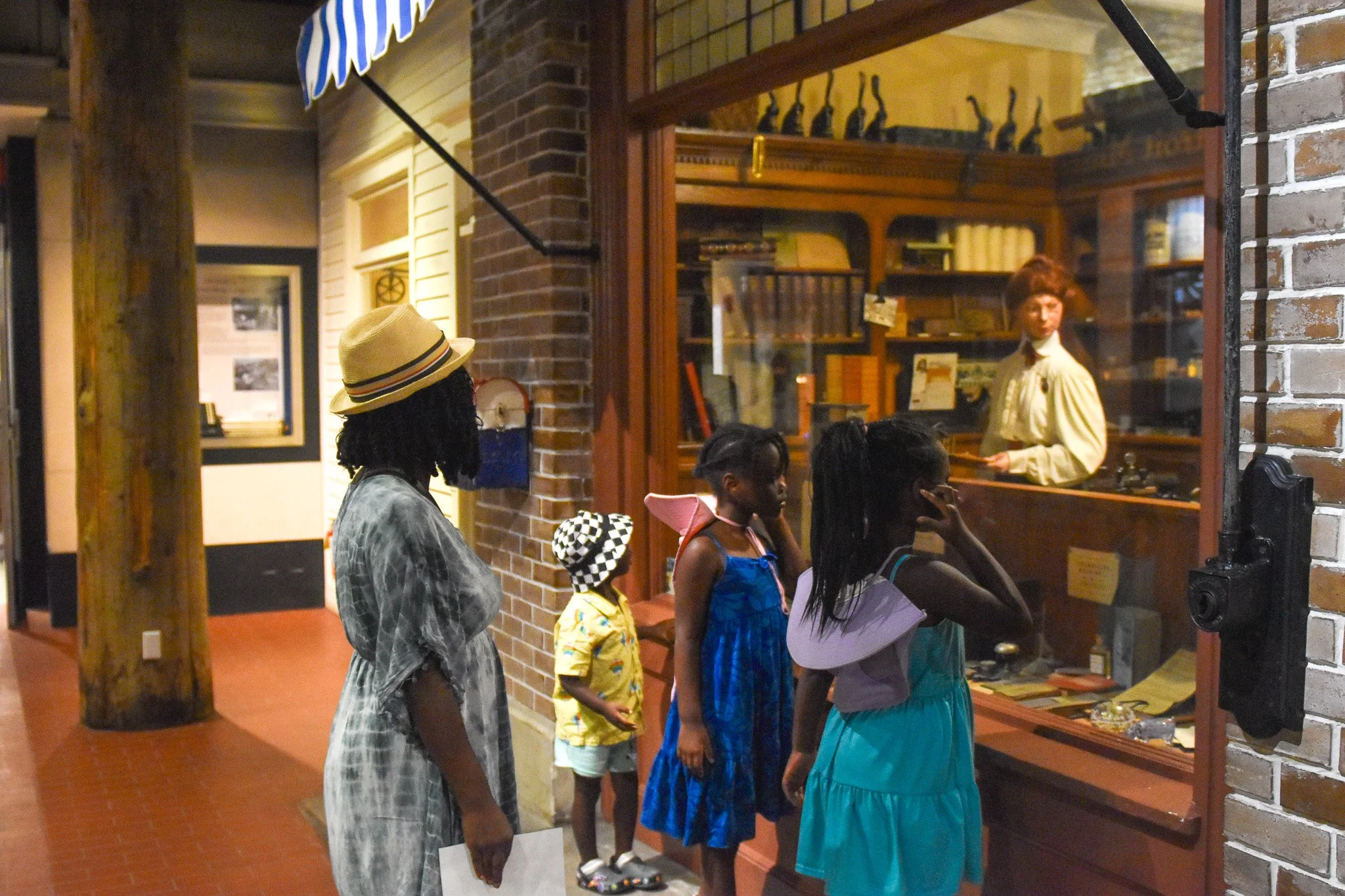 A group of children at the Detroit Historical Society, looking at a glass display case with artifacts. A woman mannequin stands behind the display case talking to the children. The museum has brick walls and shelves with various objects.