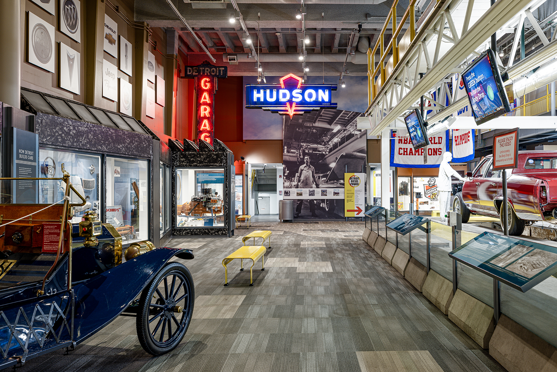 The America's Motor City exhibit at the Detroit Historical Society featuring vintage cars, framed car logos on the wall, neon signs reading 'Hudson' and 'Detroit Garage,' display cases with car parts and a model ship.