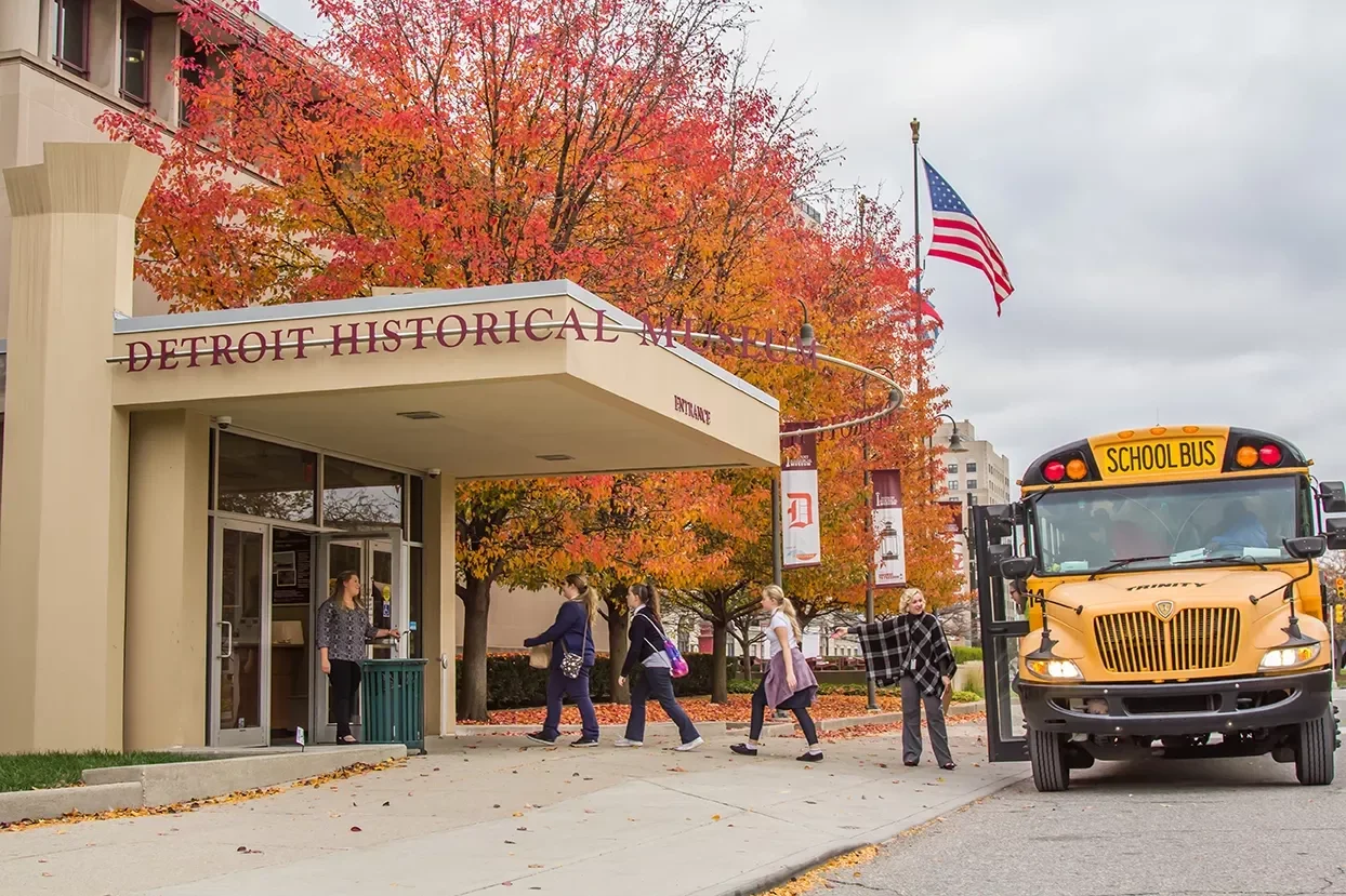 People walking off a bus into the Detroit Historical Museum during fall, with autumn leaves on trees and on the ground and an American flag flying in the background.