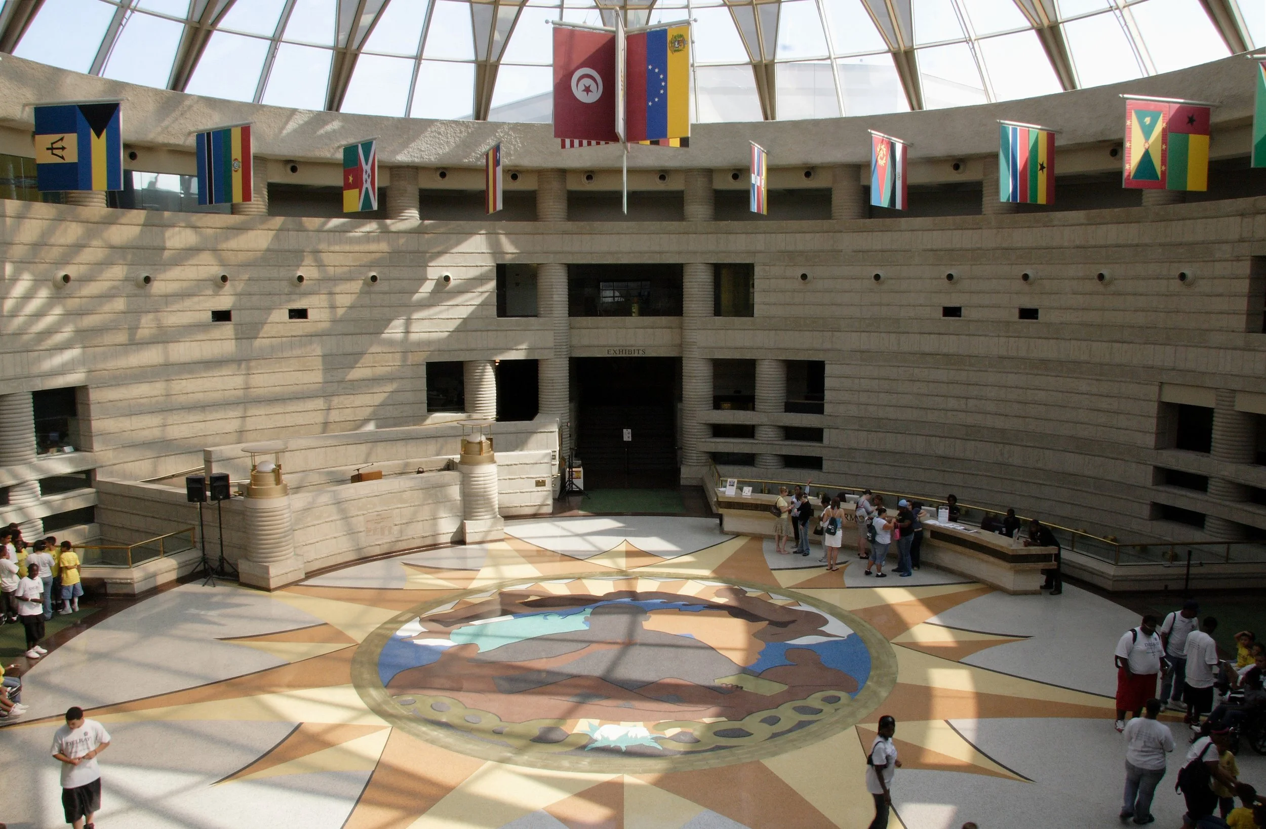 The Ring of Genealogy at the Charles H. Wright Museum. View of an indoor space with a skylight ceiling, hanging flags of various countries, and groups of people walking and standing around. The floor has a colorful, circular globe design.
