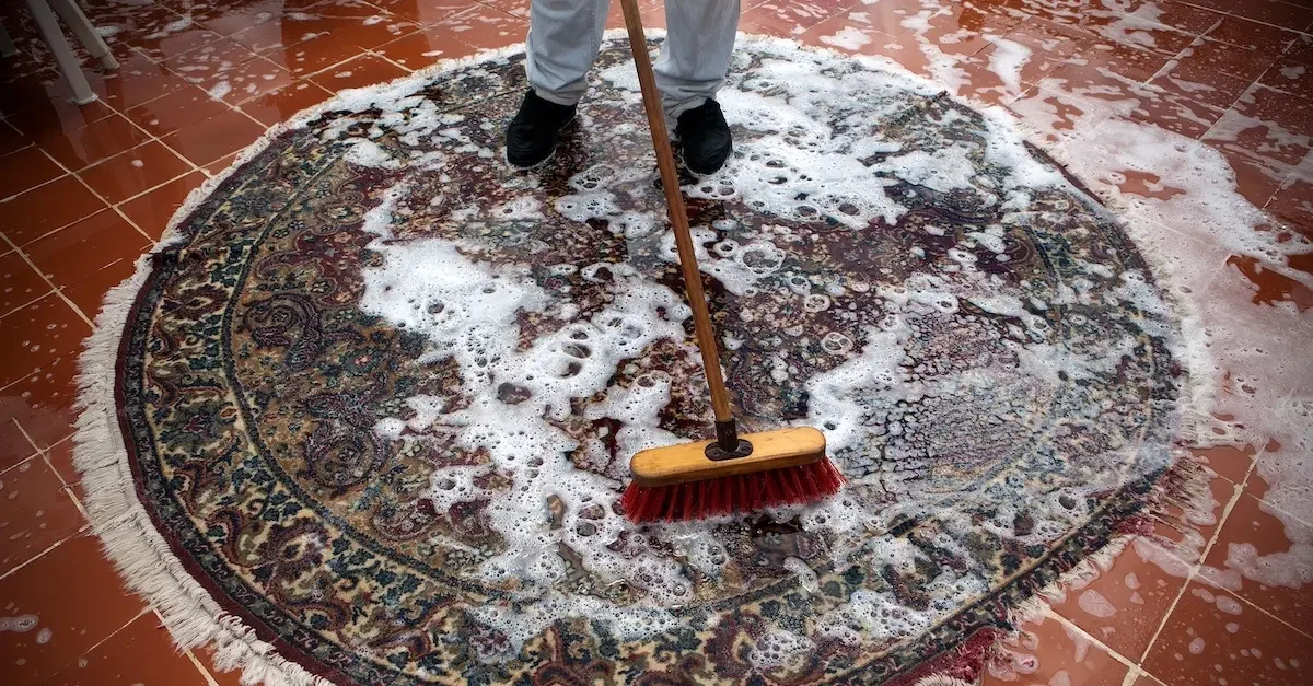 Person sweeping a colorful, patterned rug with a broom while cleaning soap suds and water from the tiled floor.