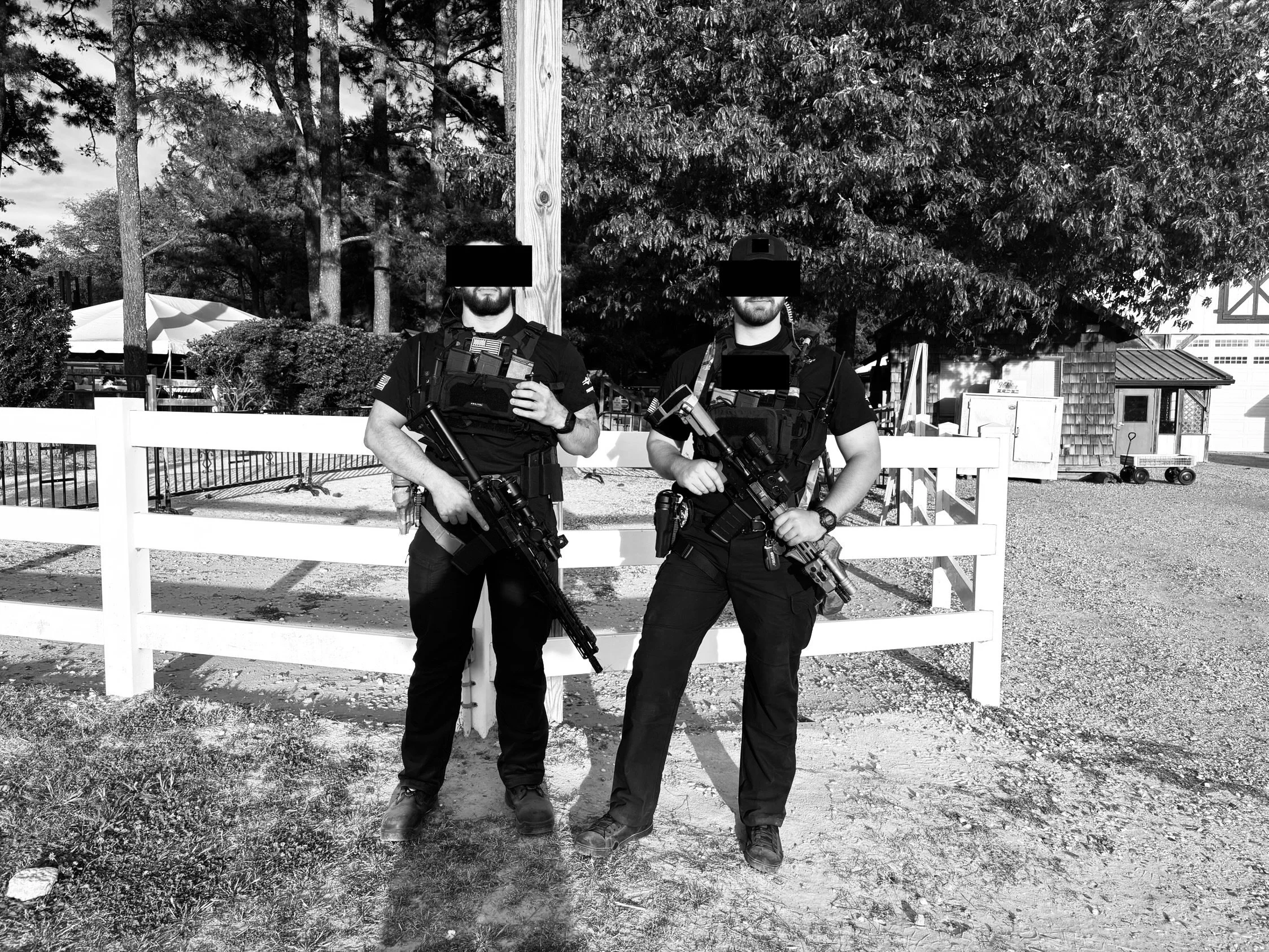 Two men dressed in tactical gear, holding rifles, standing outdoors behind a white fence, with trees and a small building in the background.