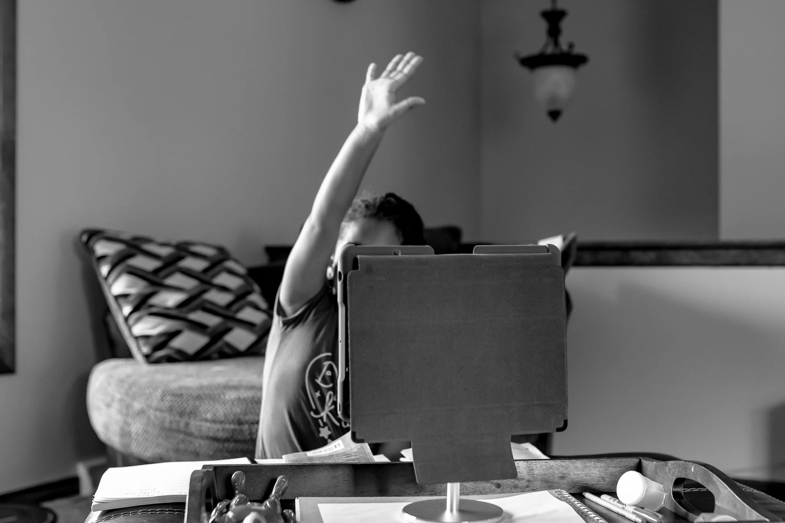 A child sitting at a table behind a tablet, raising their hand. The child's face is partially obscured, and they are in a room with a wall, a patterned chair, and a decorative light fixture in the background.
