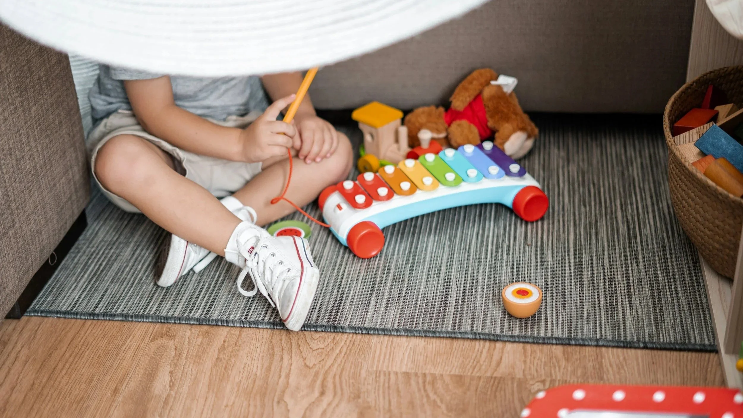 Child sitting under a blanket playing with a colorful xylophone on a gray carpeted floor.