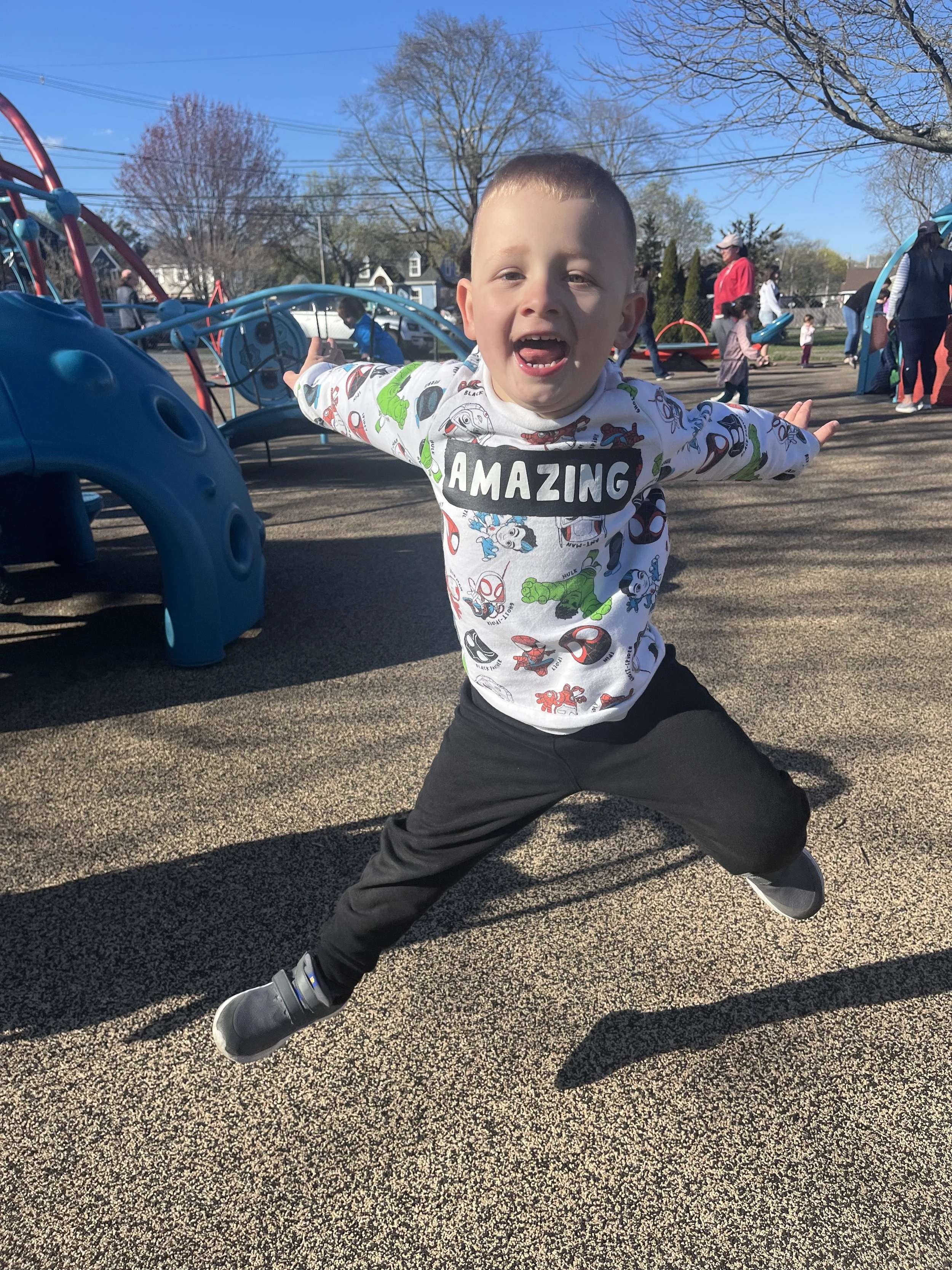 Young boy with outstretched arms, wearing a white shirt with colorful cartoon characters and the word 'AMAZING' on it, standing with legs apart on a playground with other children and adults in the background.