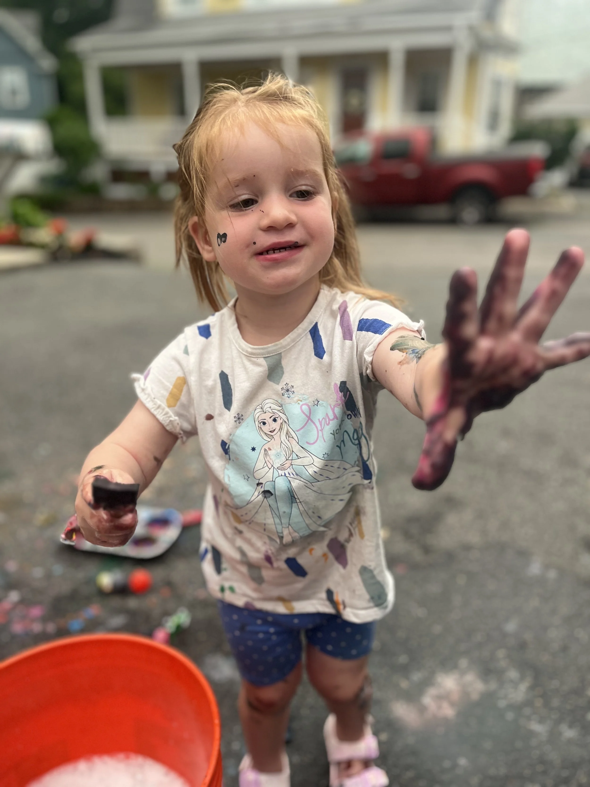 Young girl with red hair and face paint, reaching towards the camera outside on a street, surrounded by colorful objects and a red bucket in the foreground.