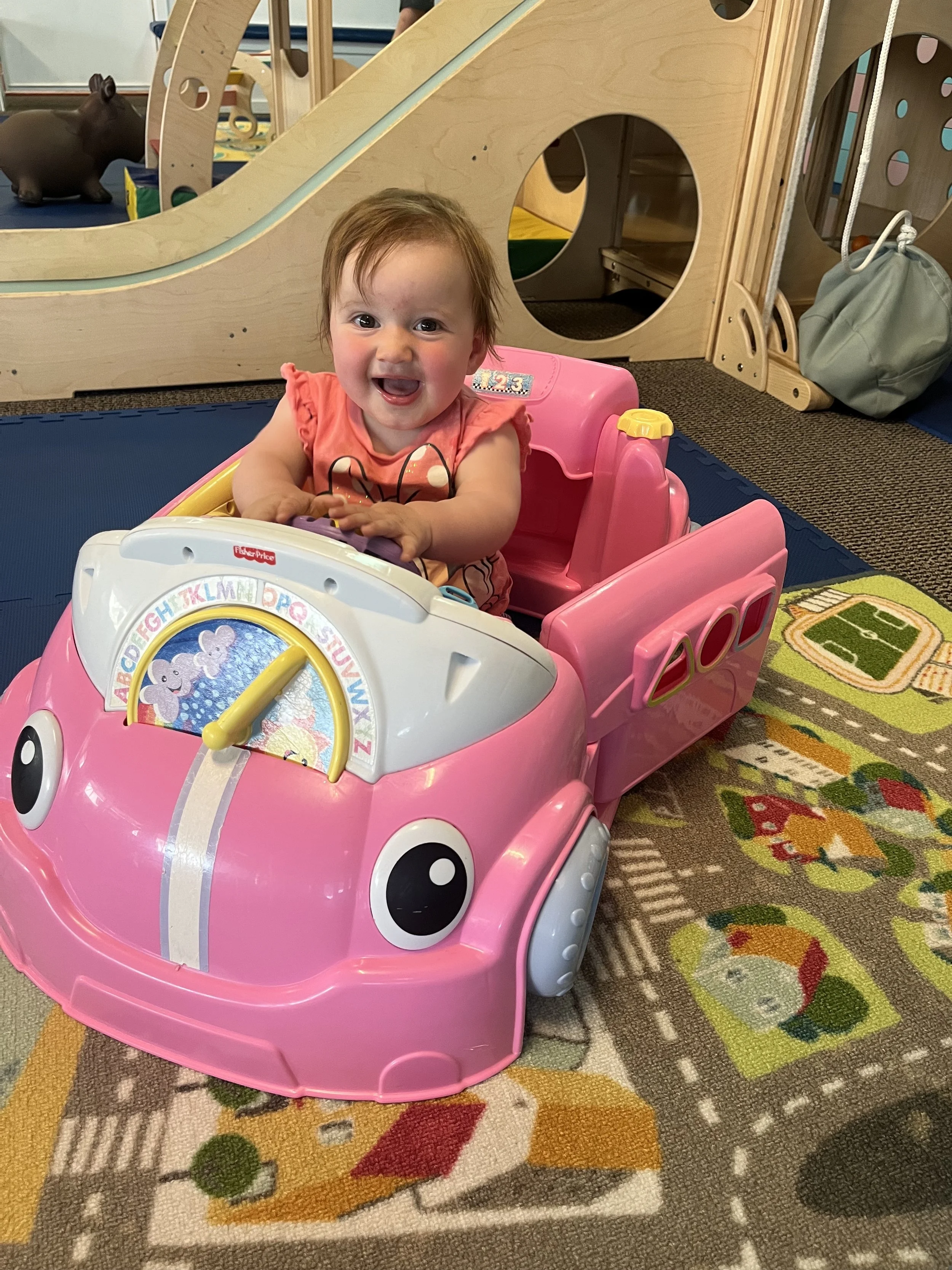 A smiling young girl with light brown hair sitting in a pink toy car with a white front panel featuring colorful alphabet letters and a picture of two elephants. She is inside an indoor play area with a colorful carpet and wooden play structures in the background.