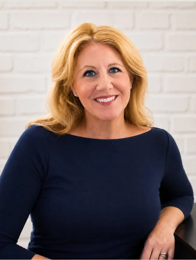 A woman with blonde hair and a navy blue top is smiling and sitting against a white brick wall.