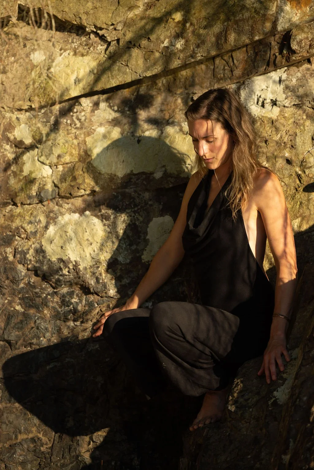 A woman with wavy brown hair sitting on rocks in a rocky outdoor setting, dressed in black, with sunlight casting shadows.
