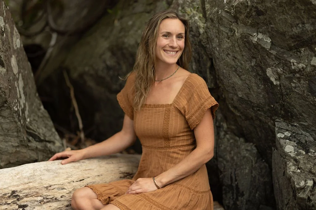A woman with long, wavy hair and a tan dress sitting on a log by rocks, smiling.